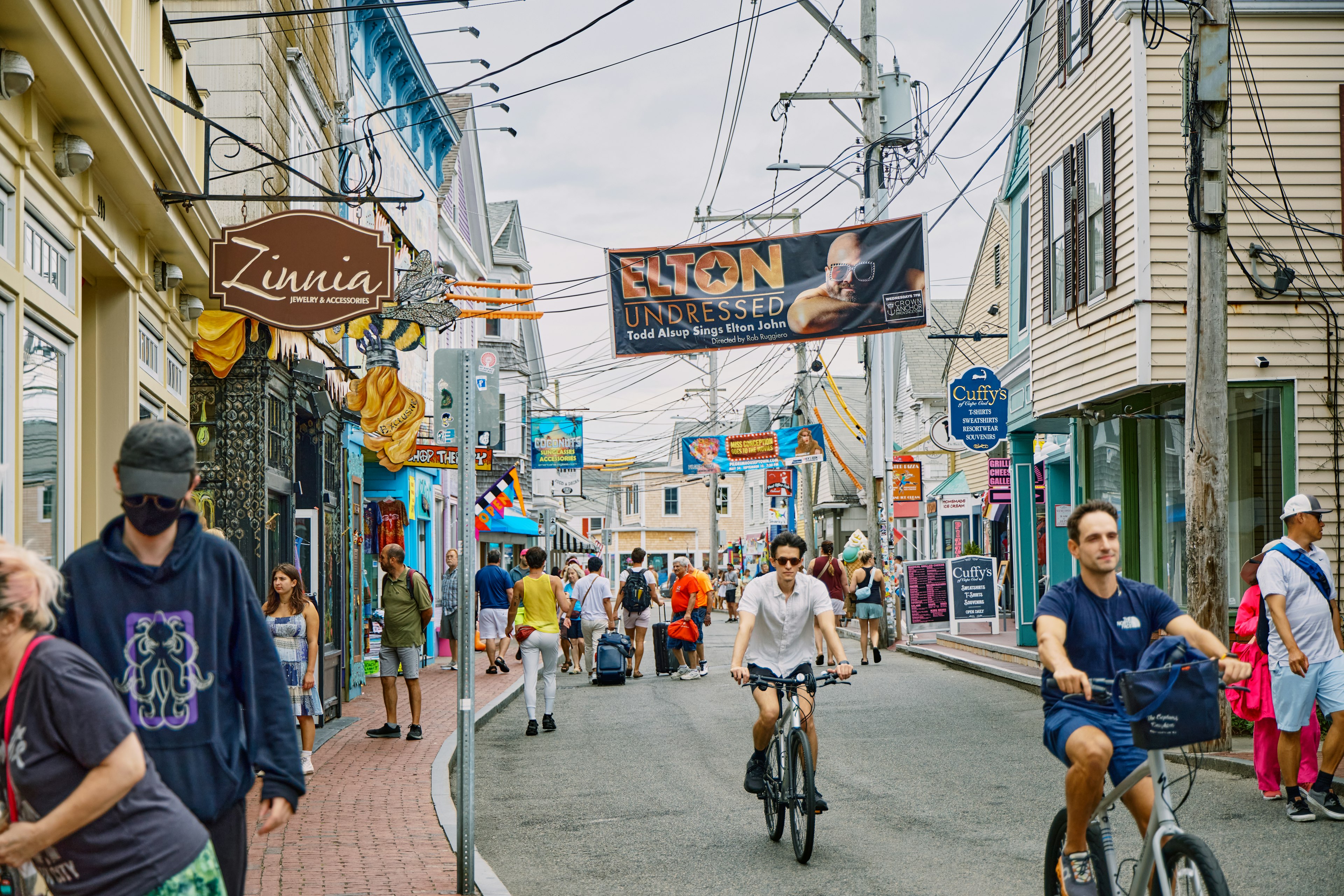 People on bikes and on foot travel down a narrow street filled with small businesses and shops with promotional banners overhead