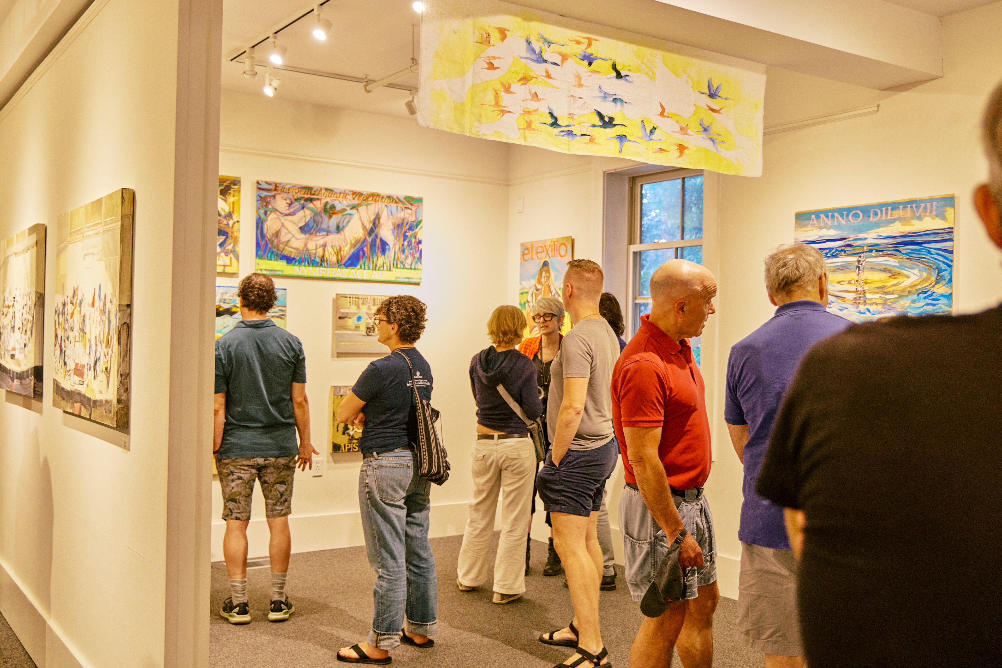 A group of people in casual summer attire admire paintings in a small art gallery