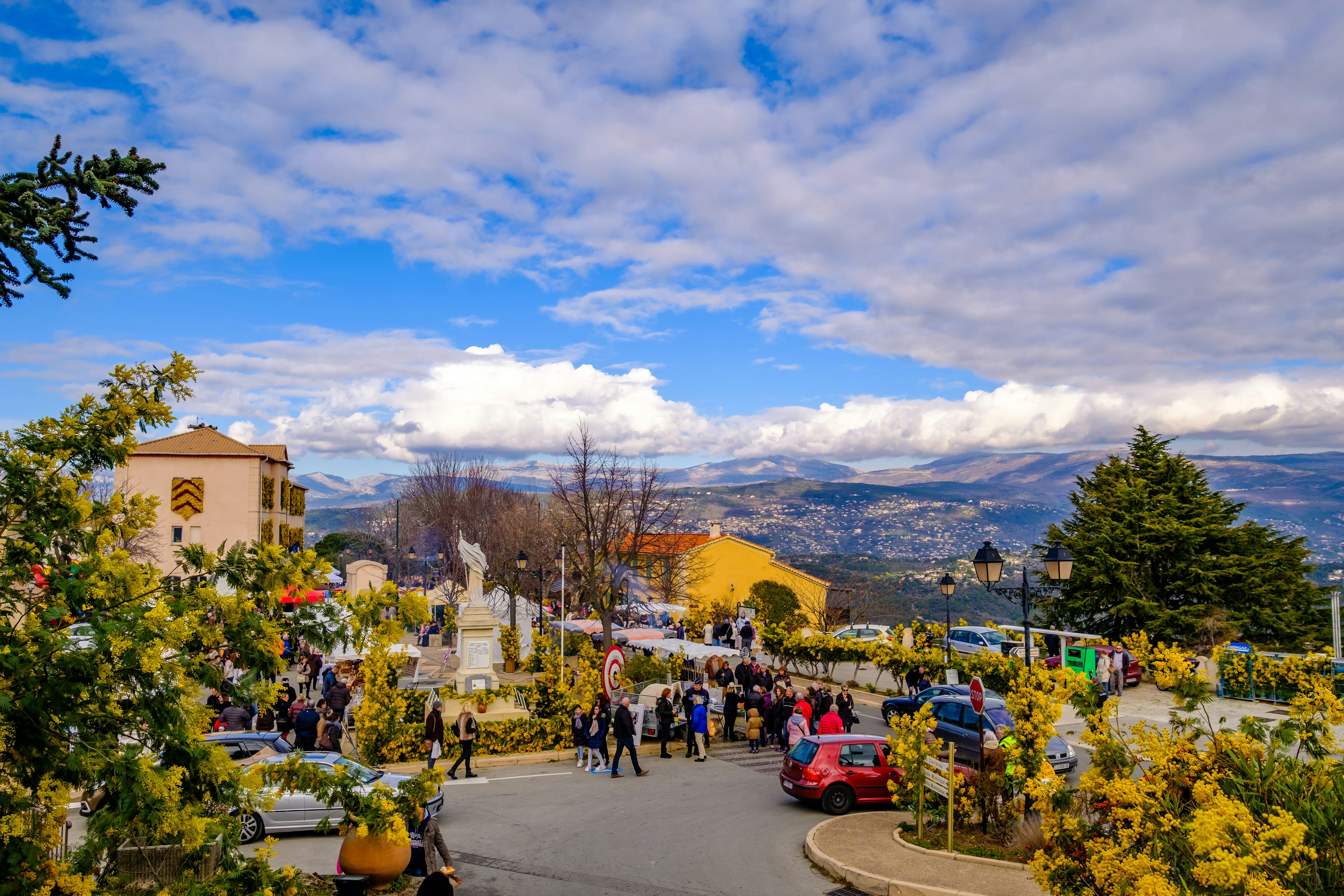 A view of a village street with people participating in a flower festival, with a beautiful view of hills and valleys opening up in the distance