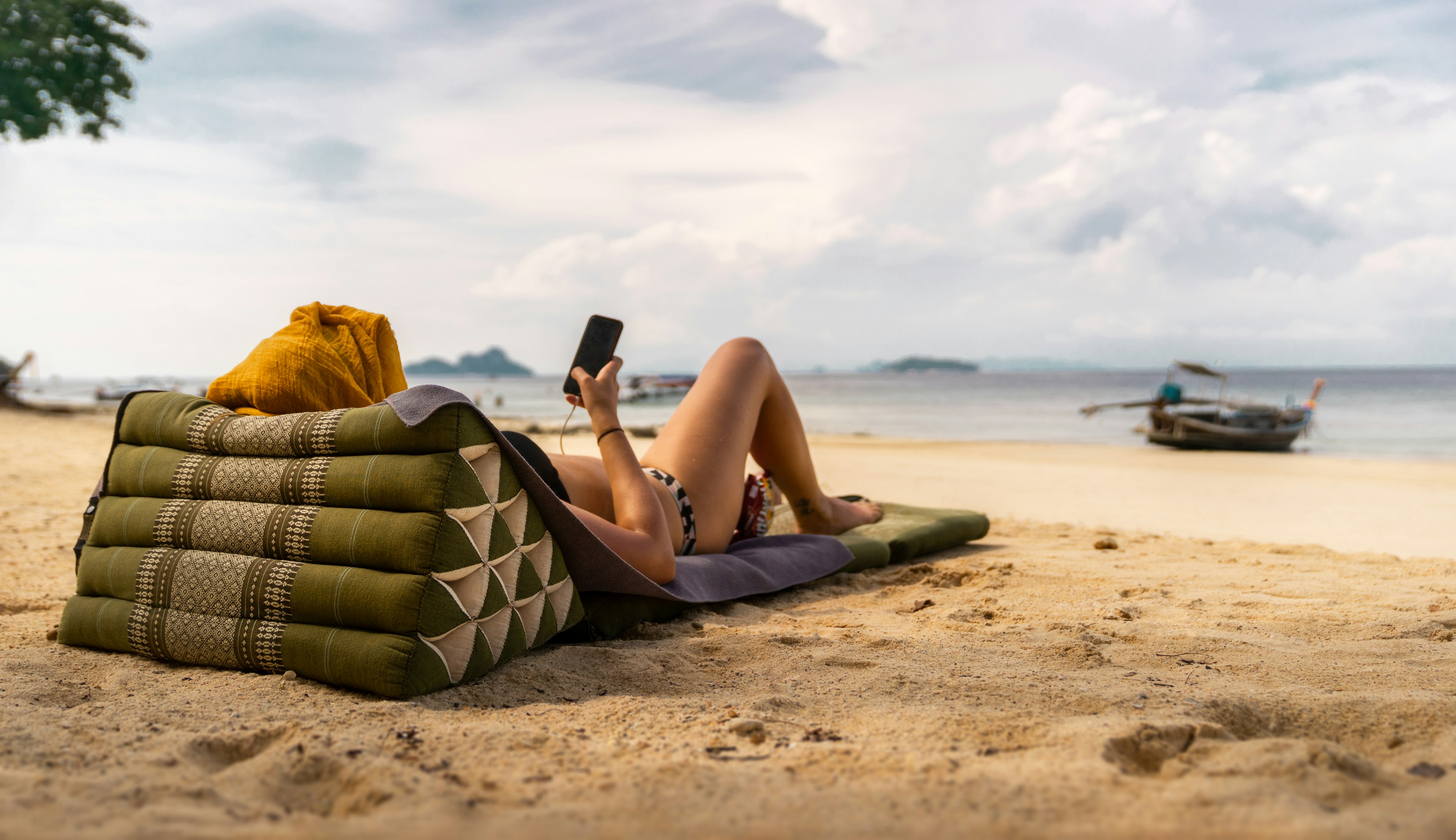 A woman lies on a triangular beach pillow on a sandy beach near a fishing boat and plays with her mobile phone