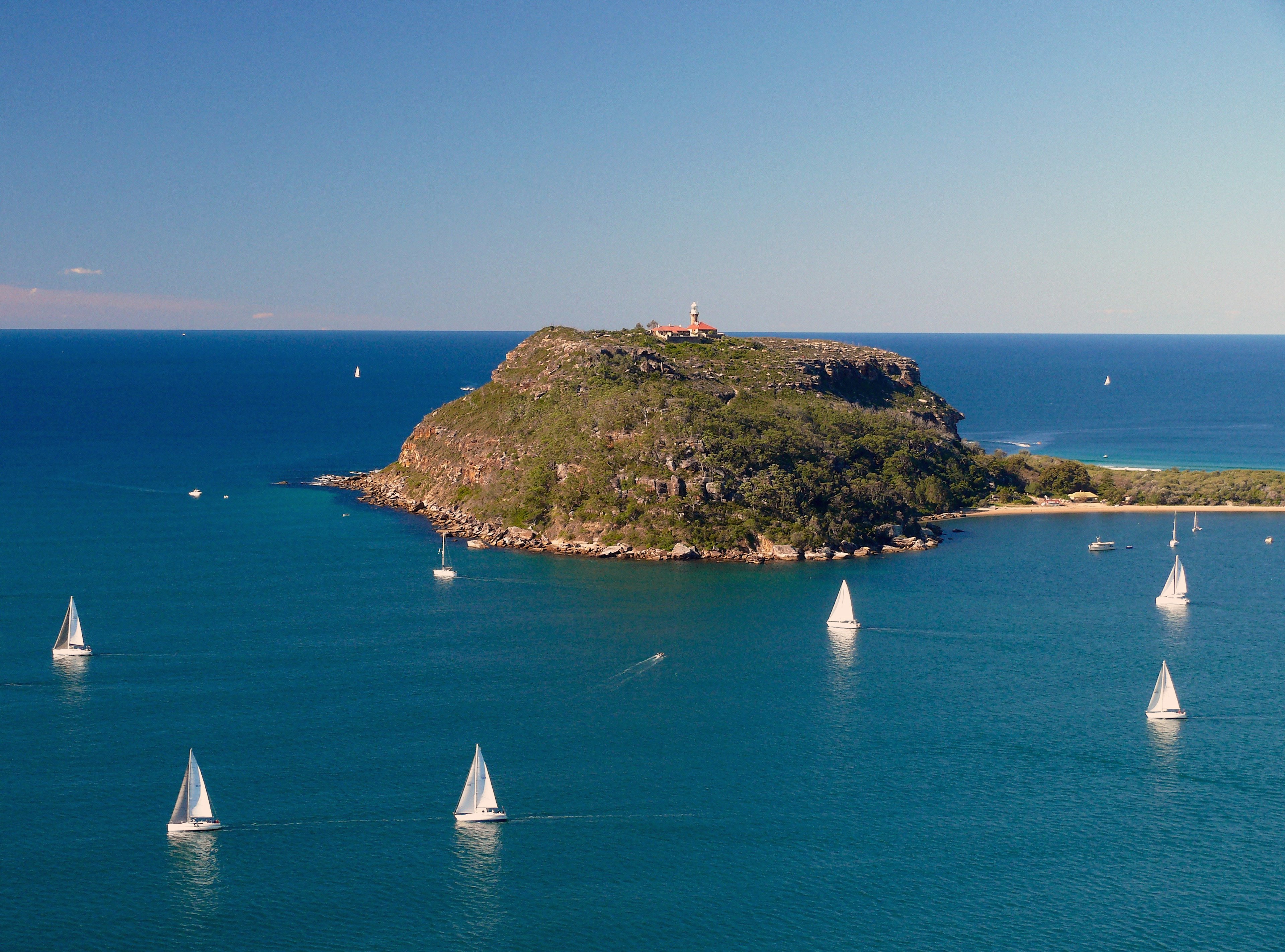 A headland topped by a lighthouse in the ocean, surrounded by boats with white sails on a sunny afternoon