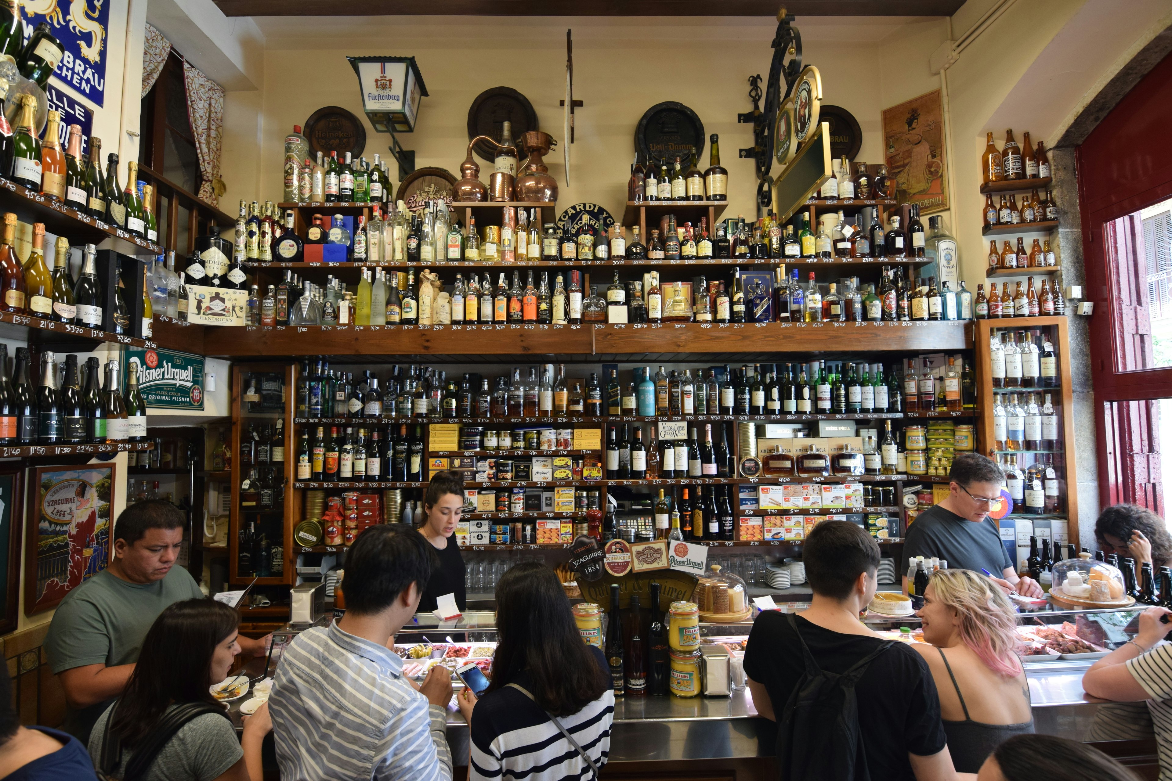 People stand a bar enjoying small plates of food and drinks. Shelves lined with wine and liquor bottles rise on the wall behind the bar.