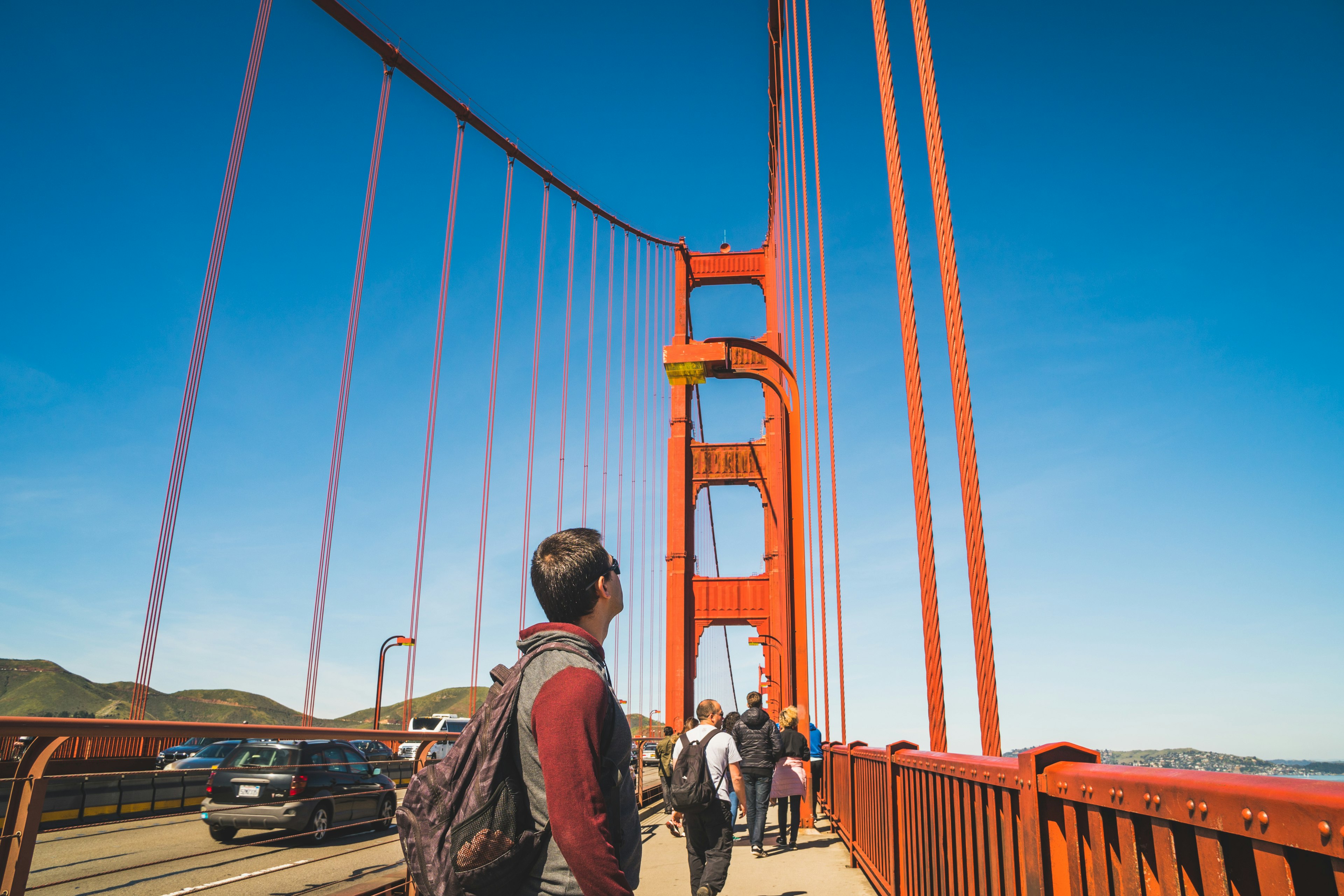 A man looks up along a pedestrian path at the side of a road on a large orange-colored suspension bridge