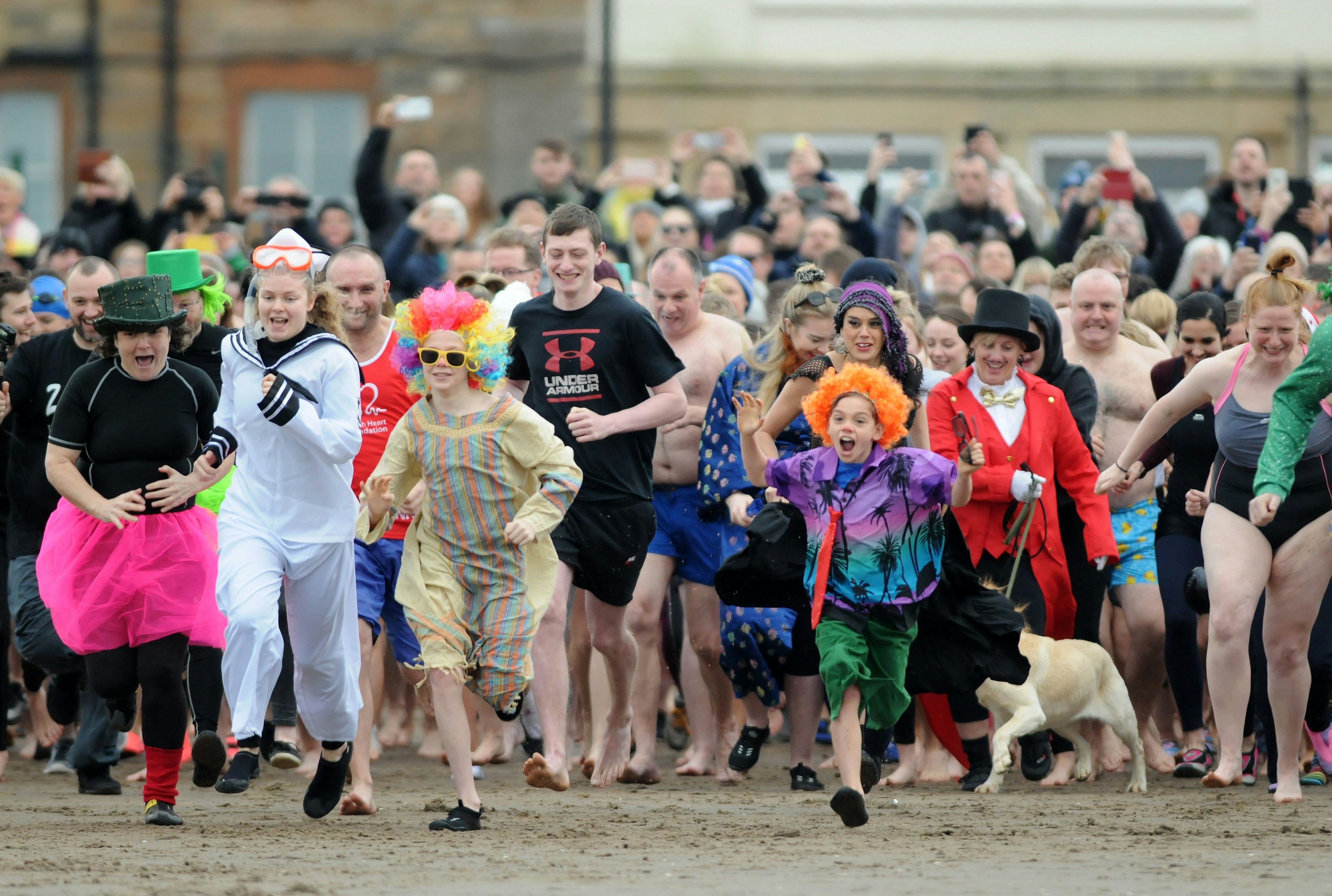 A huge crowd of people in a mix of clothes and costumes charge down a beach to run into the sea.