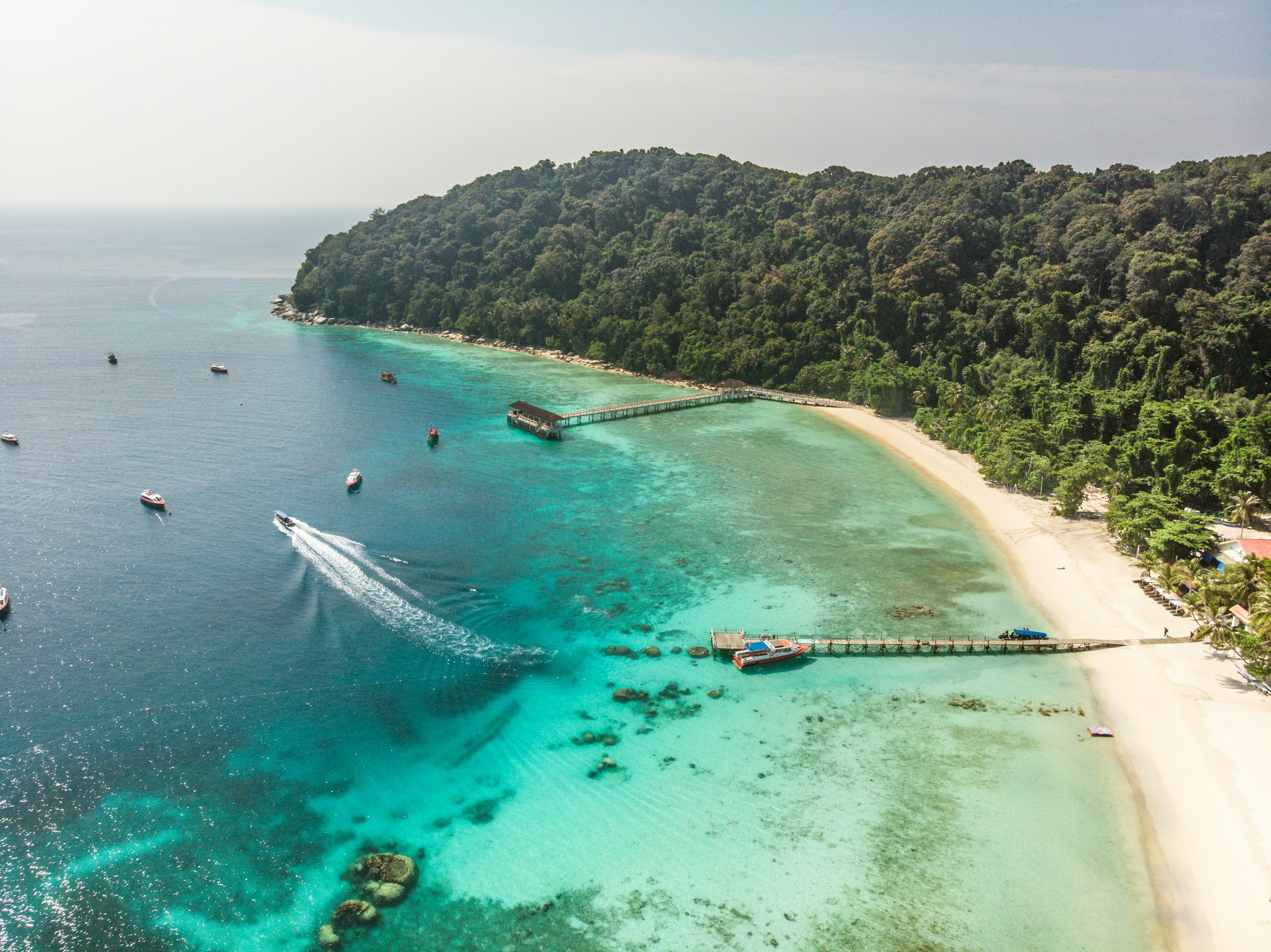 An aerial soot of boats in the clear blue waters off of Pulau Lang Tengah Island, with docks off a white-sand beach and a slope covered in green palm trees, Malaysia