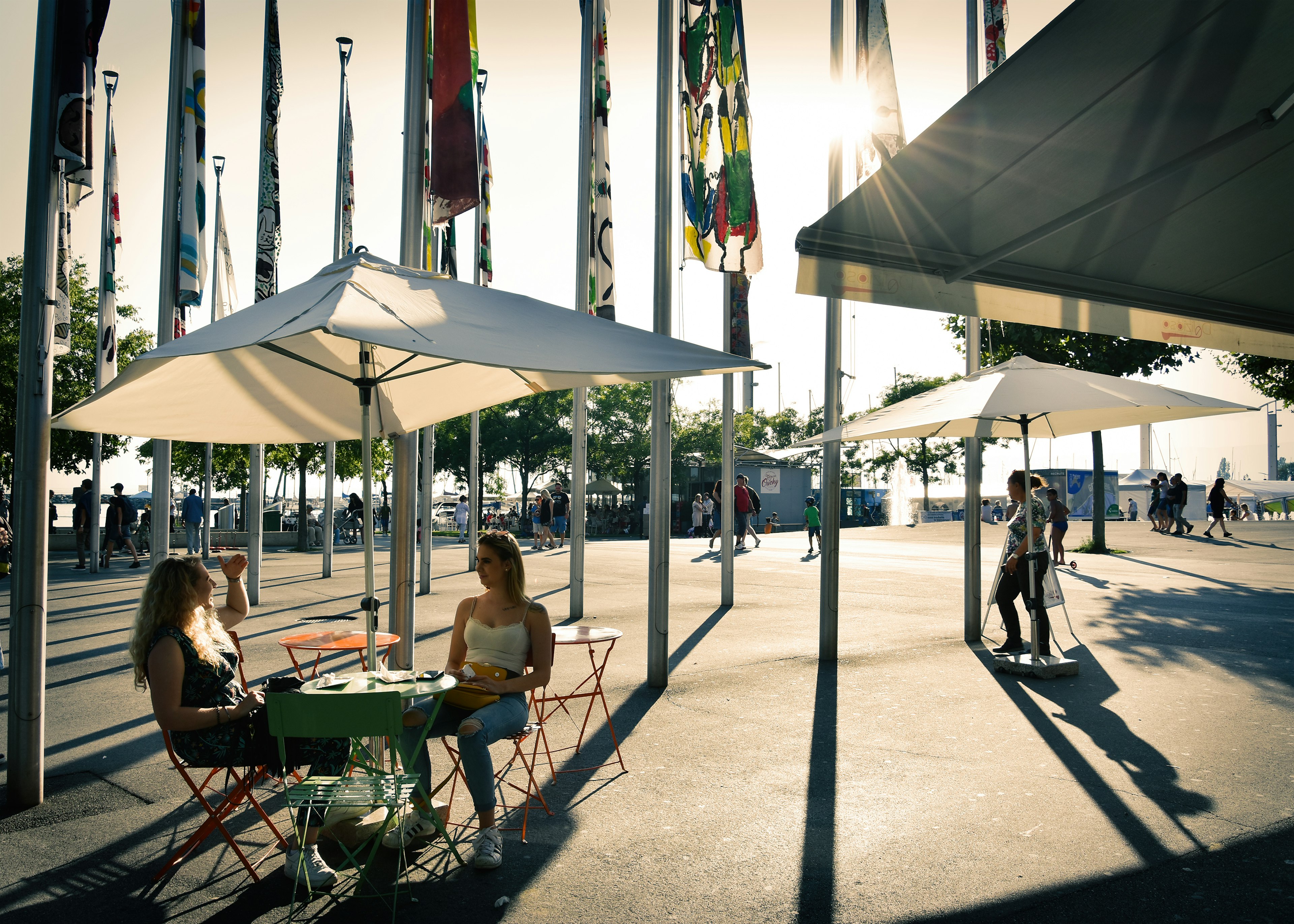 Women sit at an table outdoors under an umbrella in the slanted light, which throws shadows, of the late afternoon