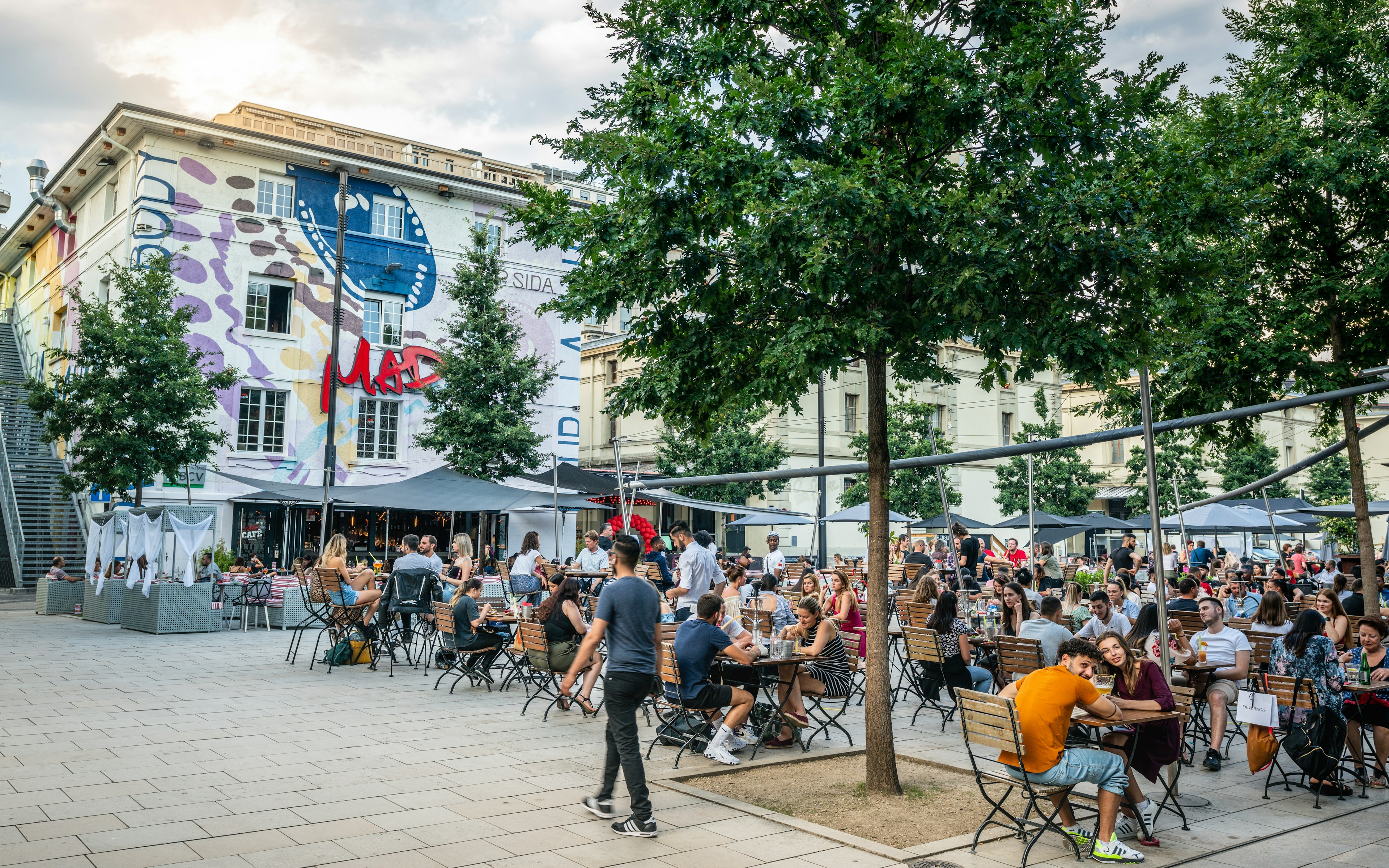 A man walks by young people seated at tables on an outdoor terrace on a city plaza in summer