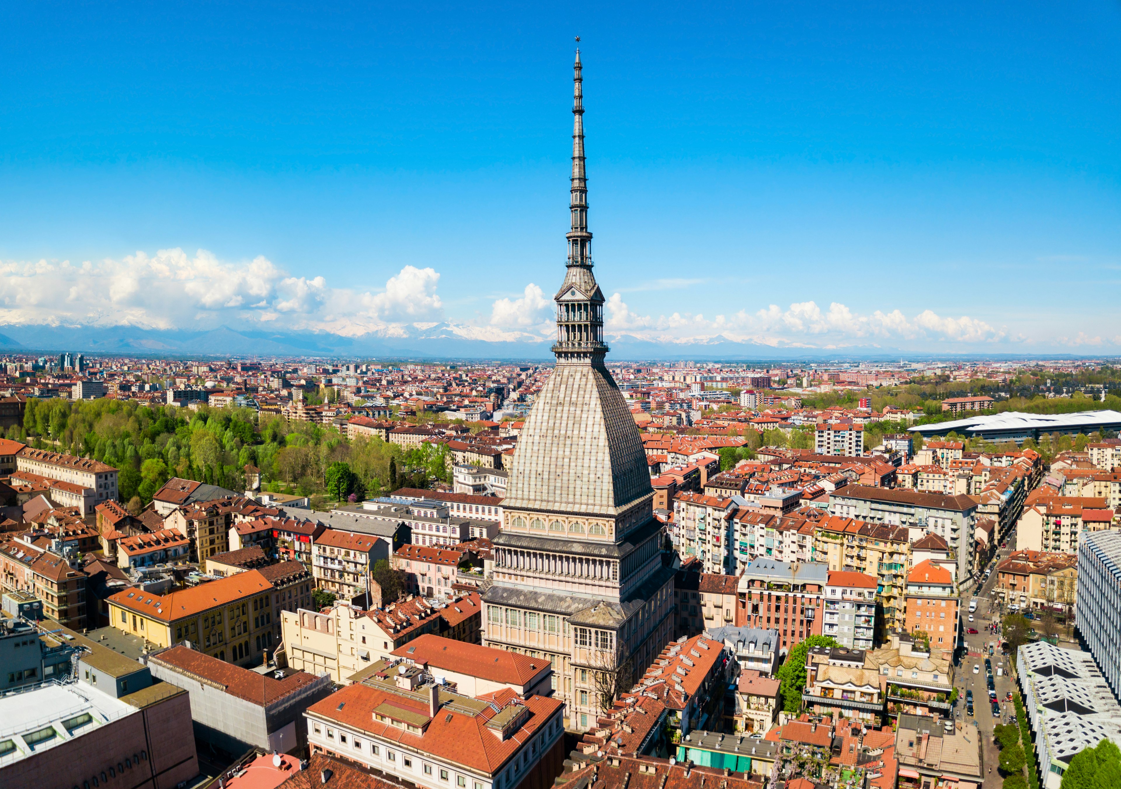 An aerial view of the Mole Antonelliana, an iconic building of the city, and the red rooftops of the central city, Turin, Italy. Snow-capped Alps are visible in the distance.