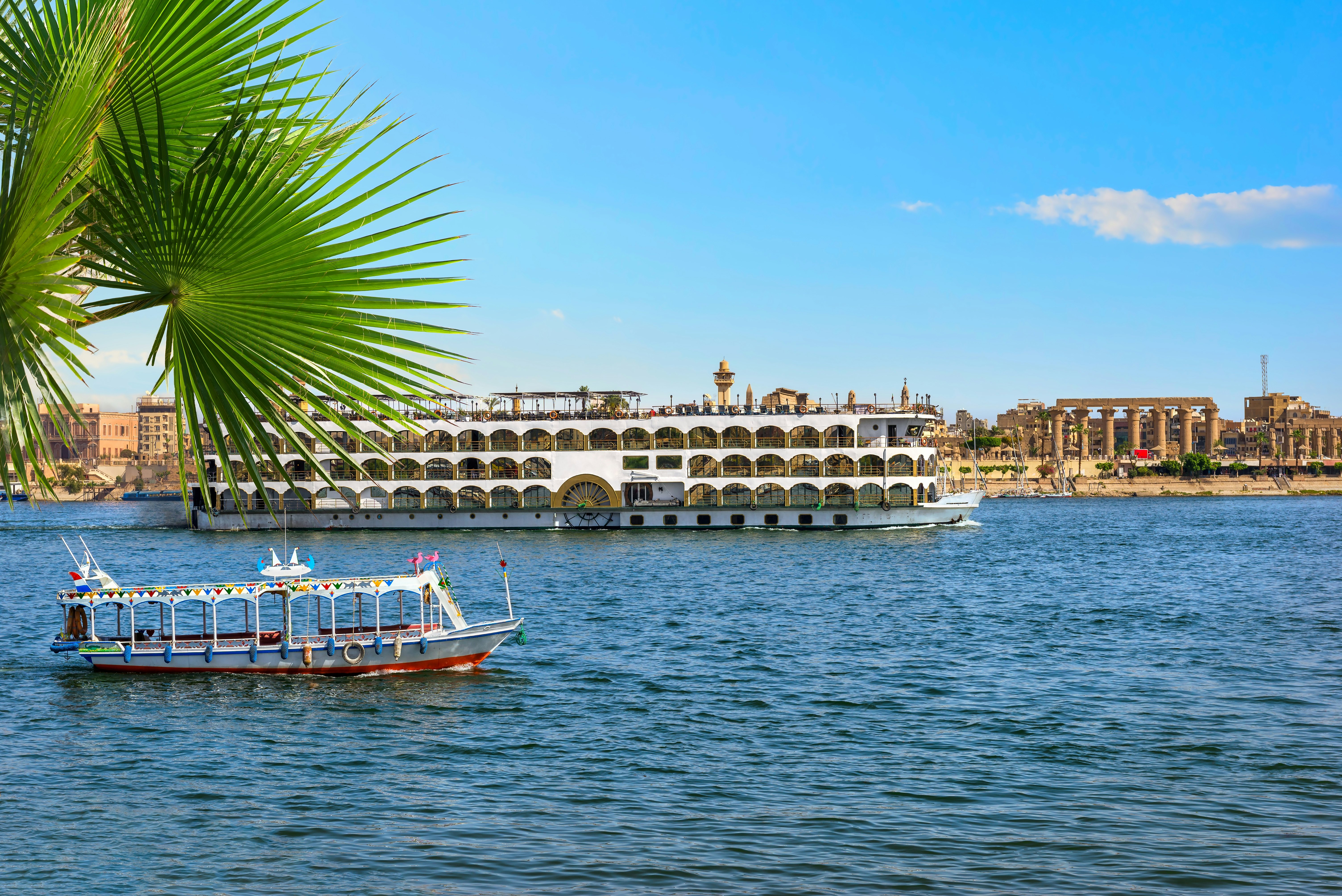 A columned temple on the riverside is obscured by a massive paddle boat with many cabins and a large sun deck. A small motor boat sails by