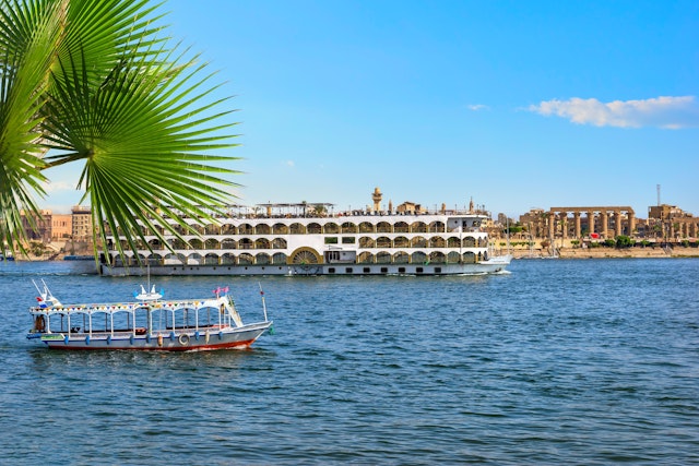 A columned temple on the riverside is obscured by a massive paddle boat with many cabins and a large sun deck. A small motor boat sails by