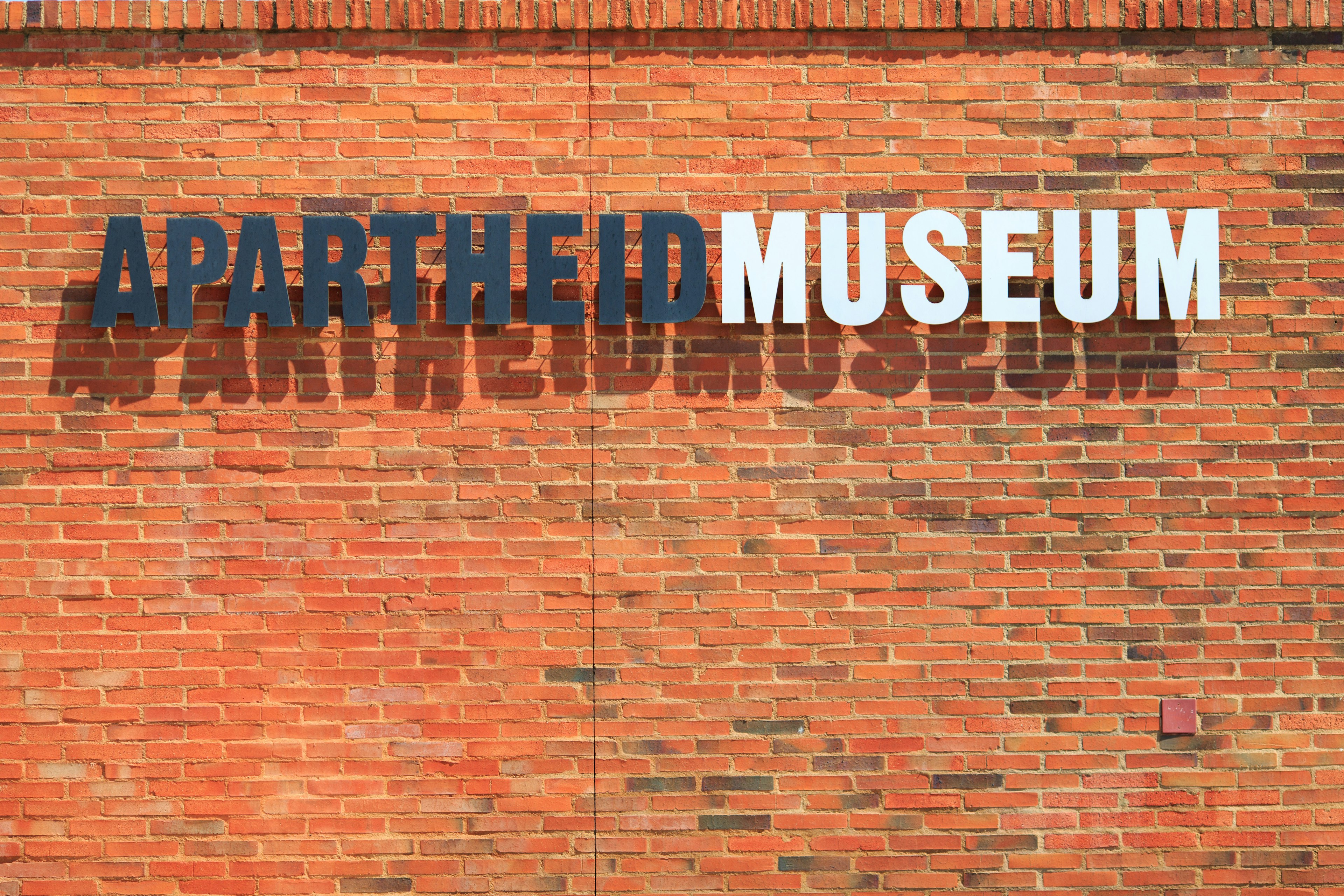 An a bright orange brick wall 'Apartheid Museum' is written in all caps. 'Apartheid' is in black, while 'Museum' is in white