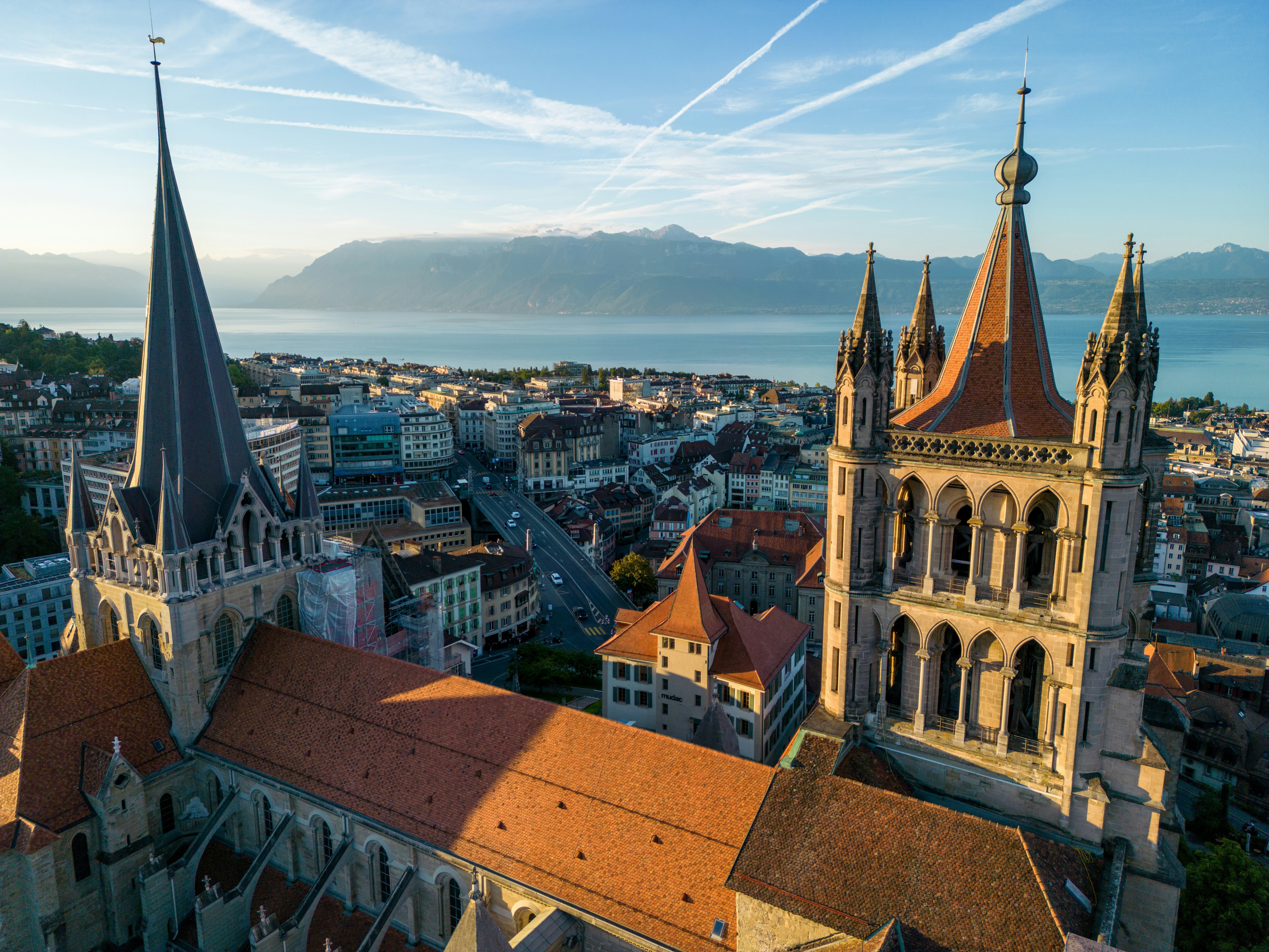 A drone aerial shot of historic cathedral towers and red rooftops with a lake and snow-capped mountains in the distance