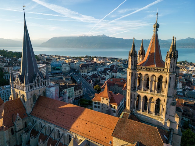 A drone aerial shot of historic cathedral towers and red rooftops with a lake and snow-capped mountains in the distance