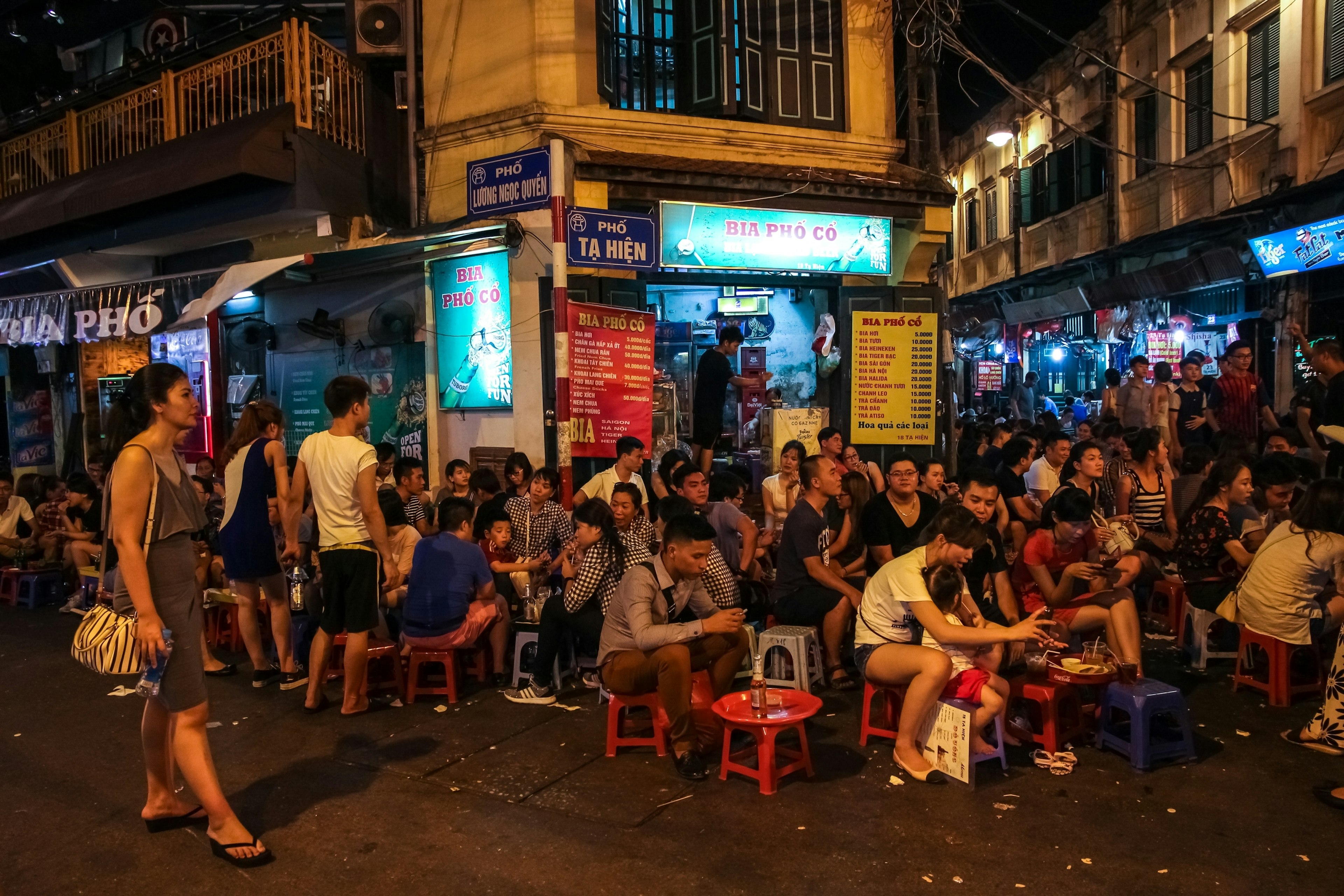 A crowded street corner in the old quarter of the city of Hanoi with young people sitting on stools while enjoying bia hoi (fresh beer).