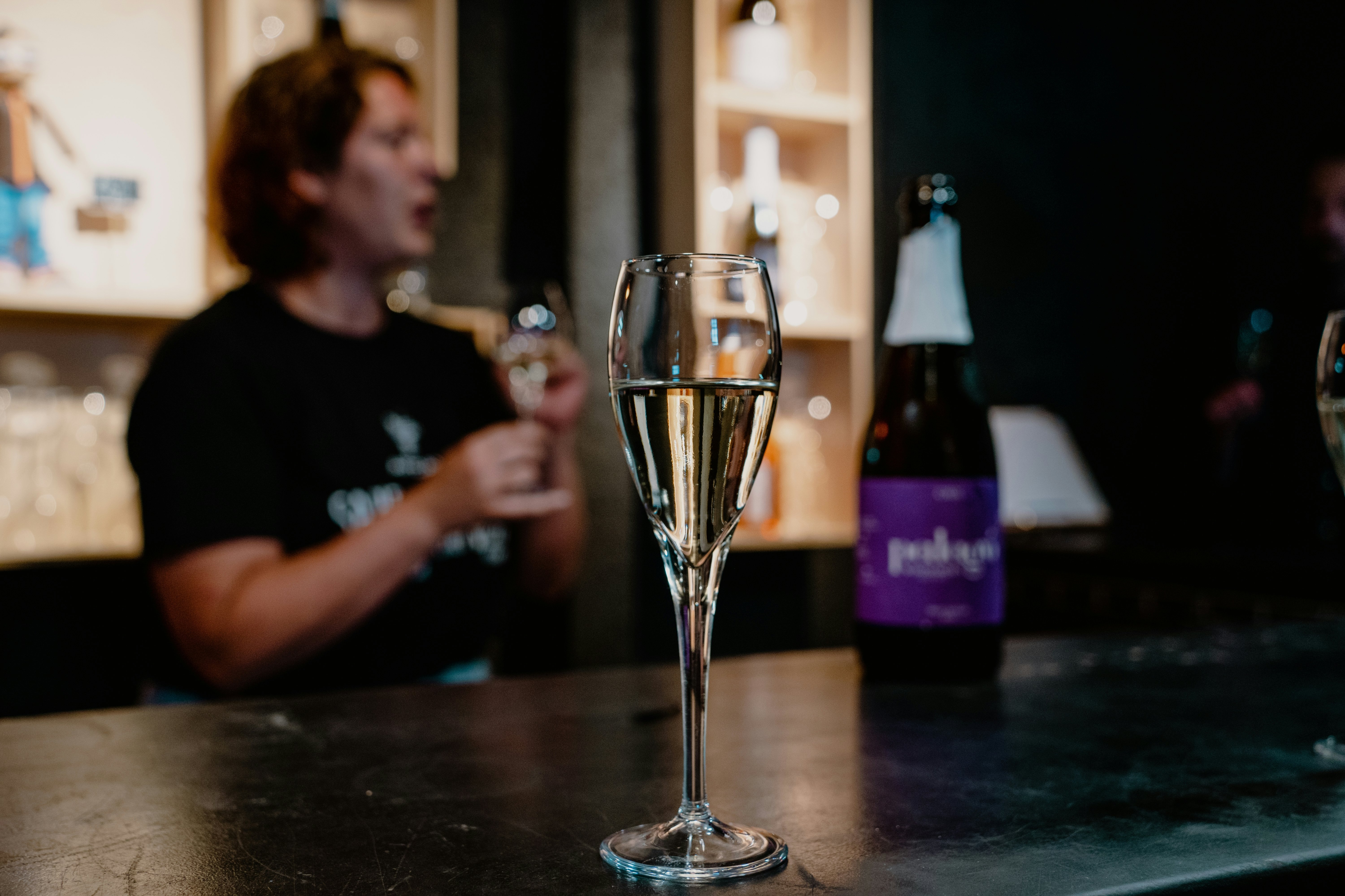 Sharp image of a flute of wine in the foreground on a black countertop, with a bottle at the right of the photo and a person standing in the background holding a flute