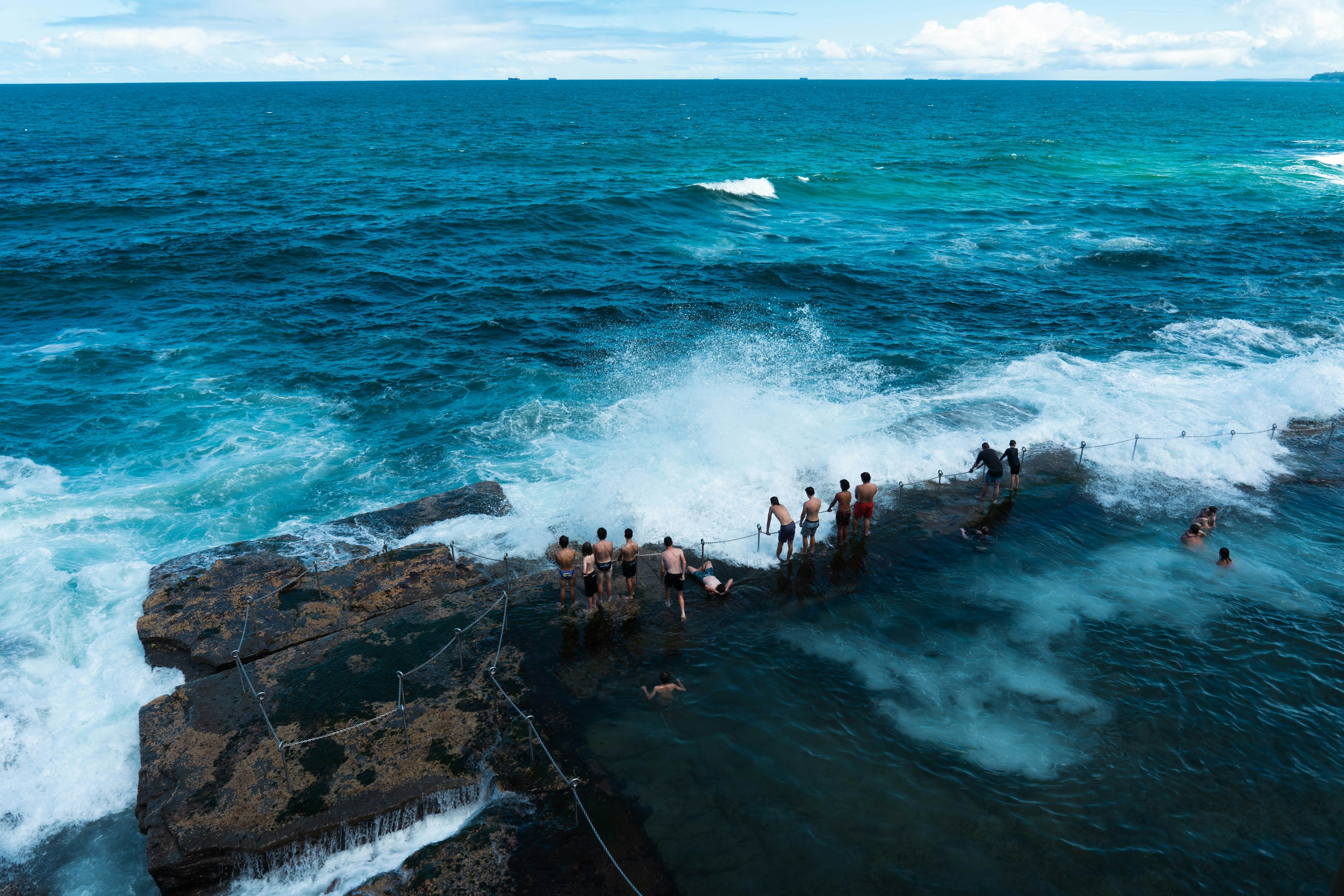 People stand on the rim of an ocean bath carved into the rock cliffs, as waves from the Pacific Ocean crash over them, Newcastle, New South Wales, Australia