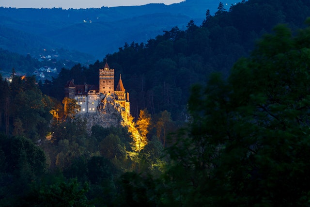 A view of a castle on a cliff top illuminated by night among shadowy hillsides