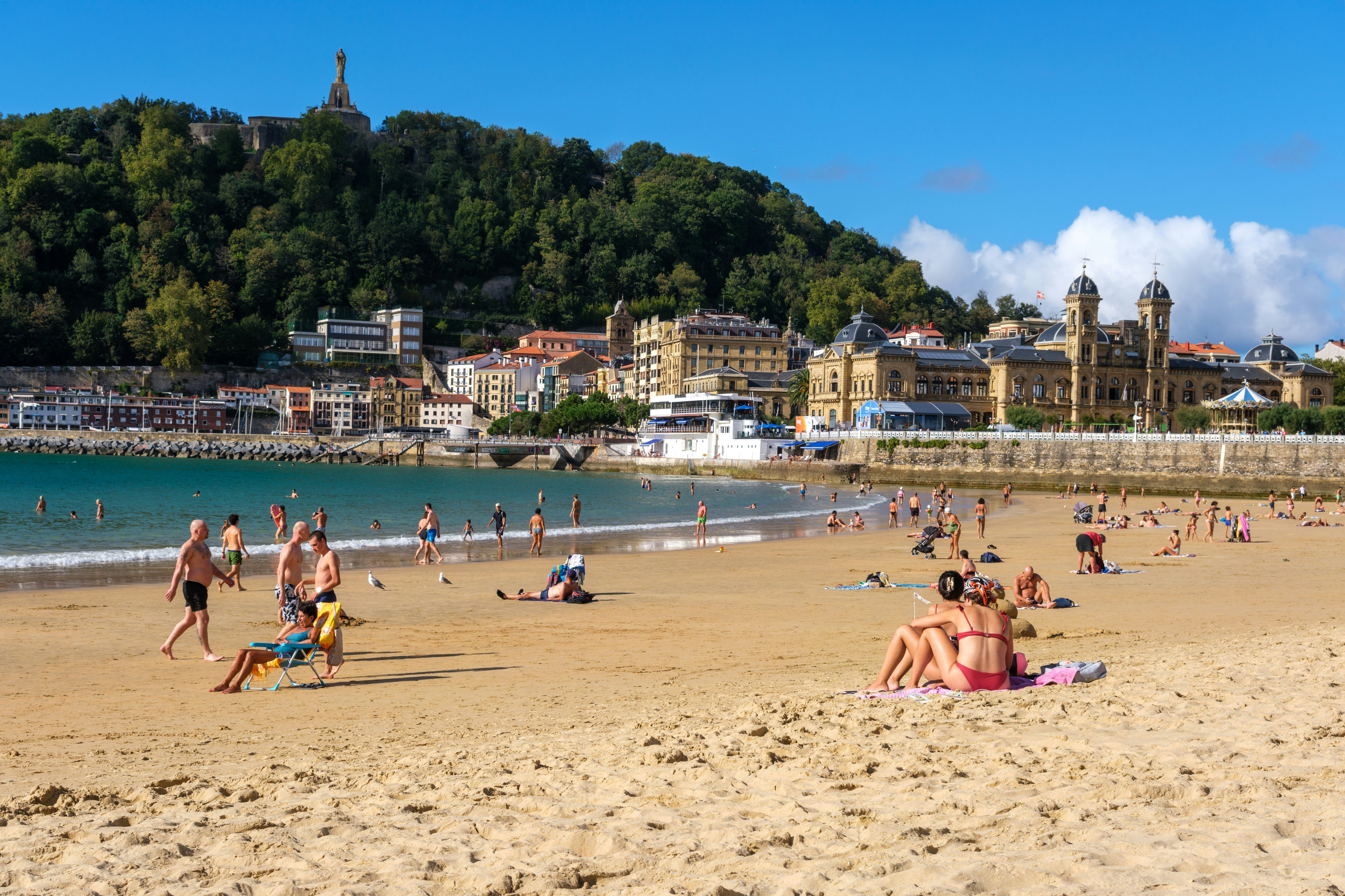 People on a sandy beach at the edge of a city tucked below a steep hill.