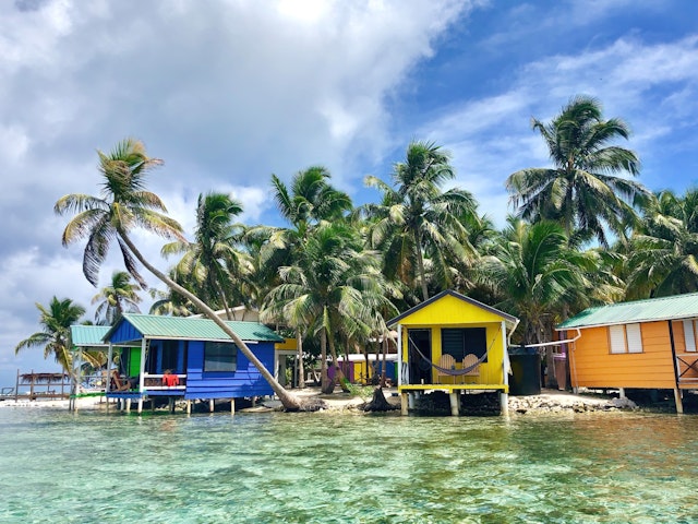 Colorful, small bungalows are built out over the clear water with palm trees behind them, Tobacco Caye, Belize