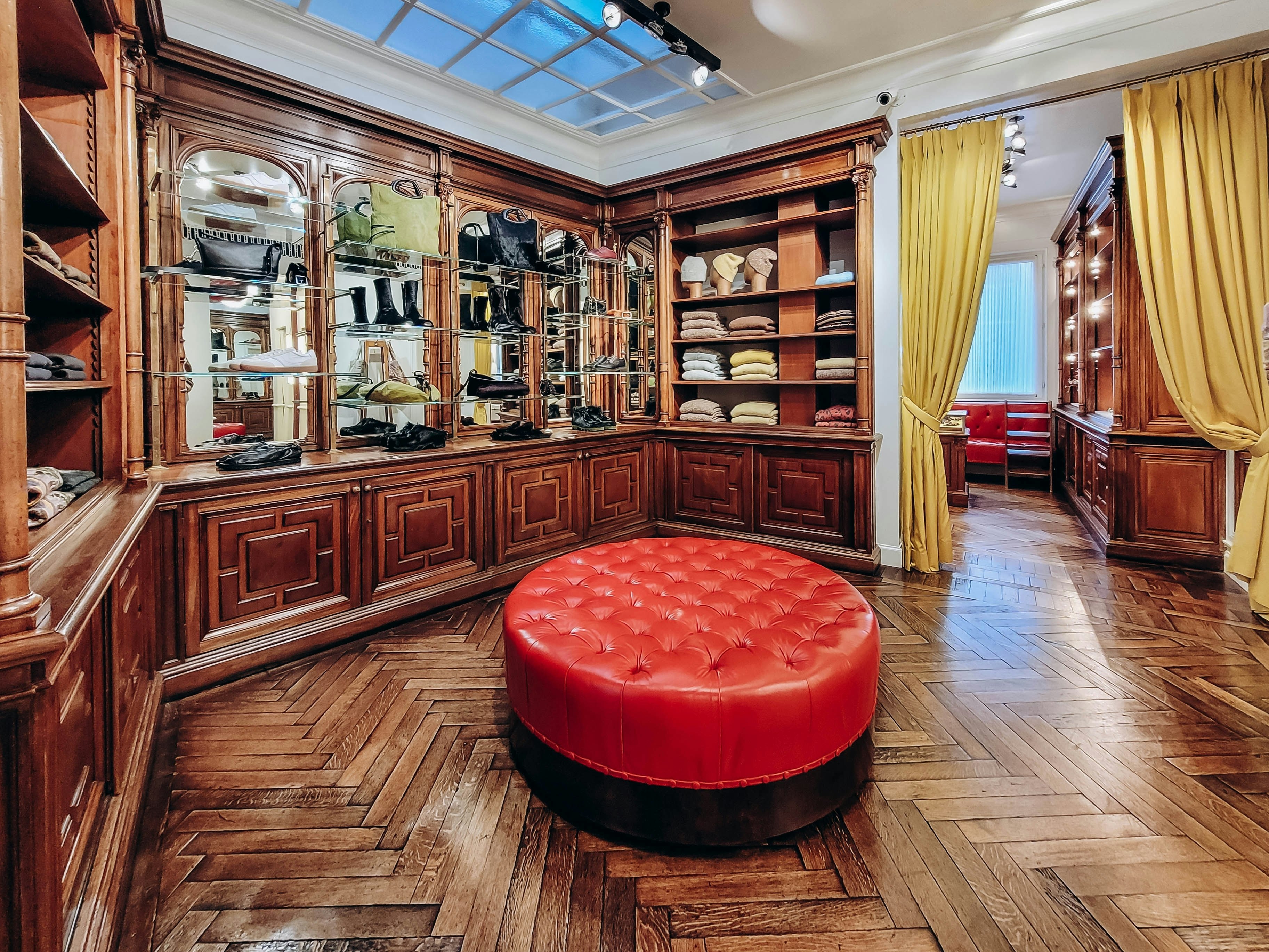 A large wood-panelled room in a department store selling shoes, clothes and bags, with a round red leather sofa in the center.