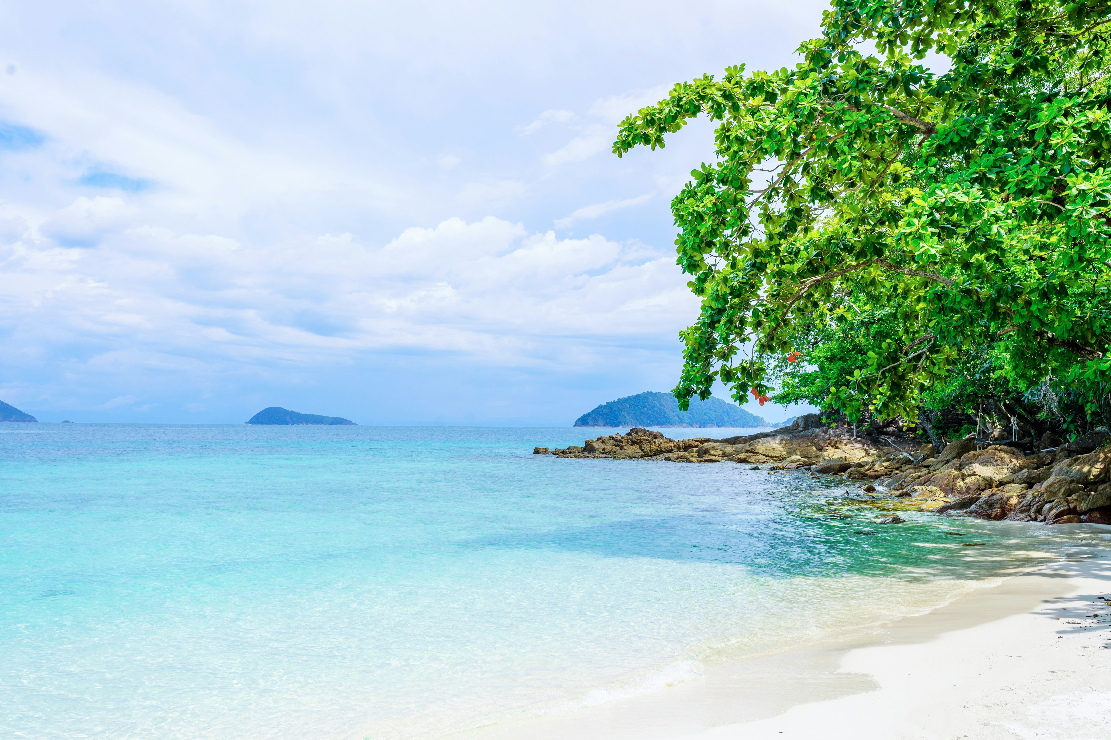 Trees grow over a white-sand beach, with islands visible in the distance on the blue water