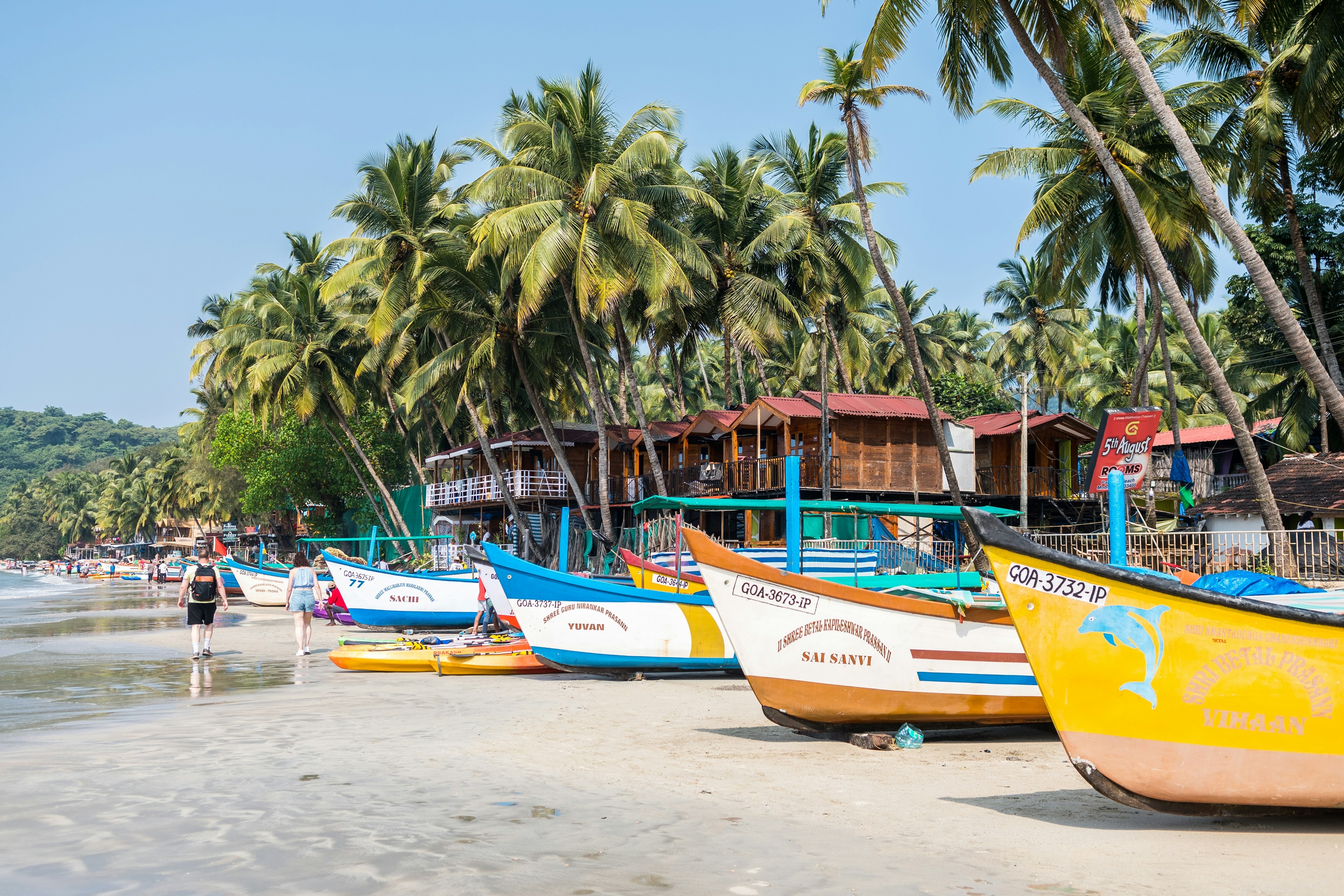 People walk by brightly colored fishing boats on the sand underneath palm trees on a beach in Goa, India