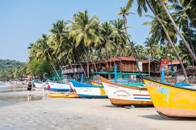 People walk by brightly colored fishing boats on the sand underneath palm trees on a beach in Goa, India