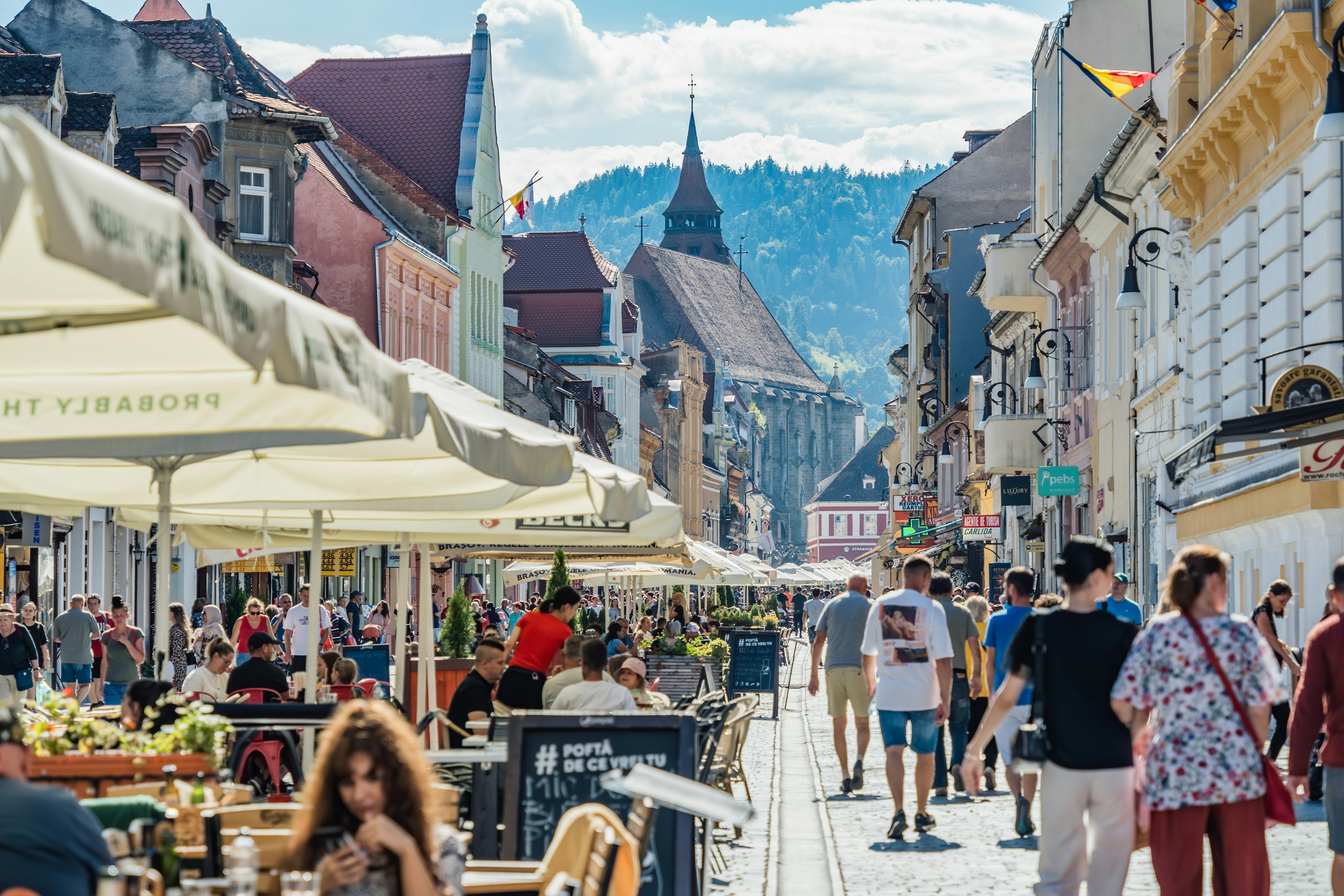 People stroll down a pedestrianized street past other people sitting at cafe tables in the historic center of Brașov, Romania. A church is visible in the distance