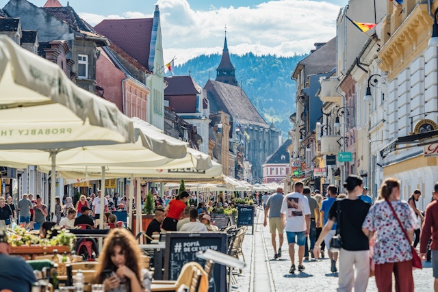 People stroll down a pedestrianized street past other people sitting at cafe tables in the historic center of Brașov, Romania. A church is visible in the distance