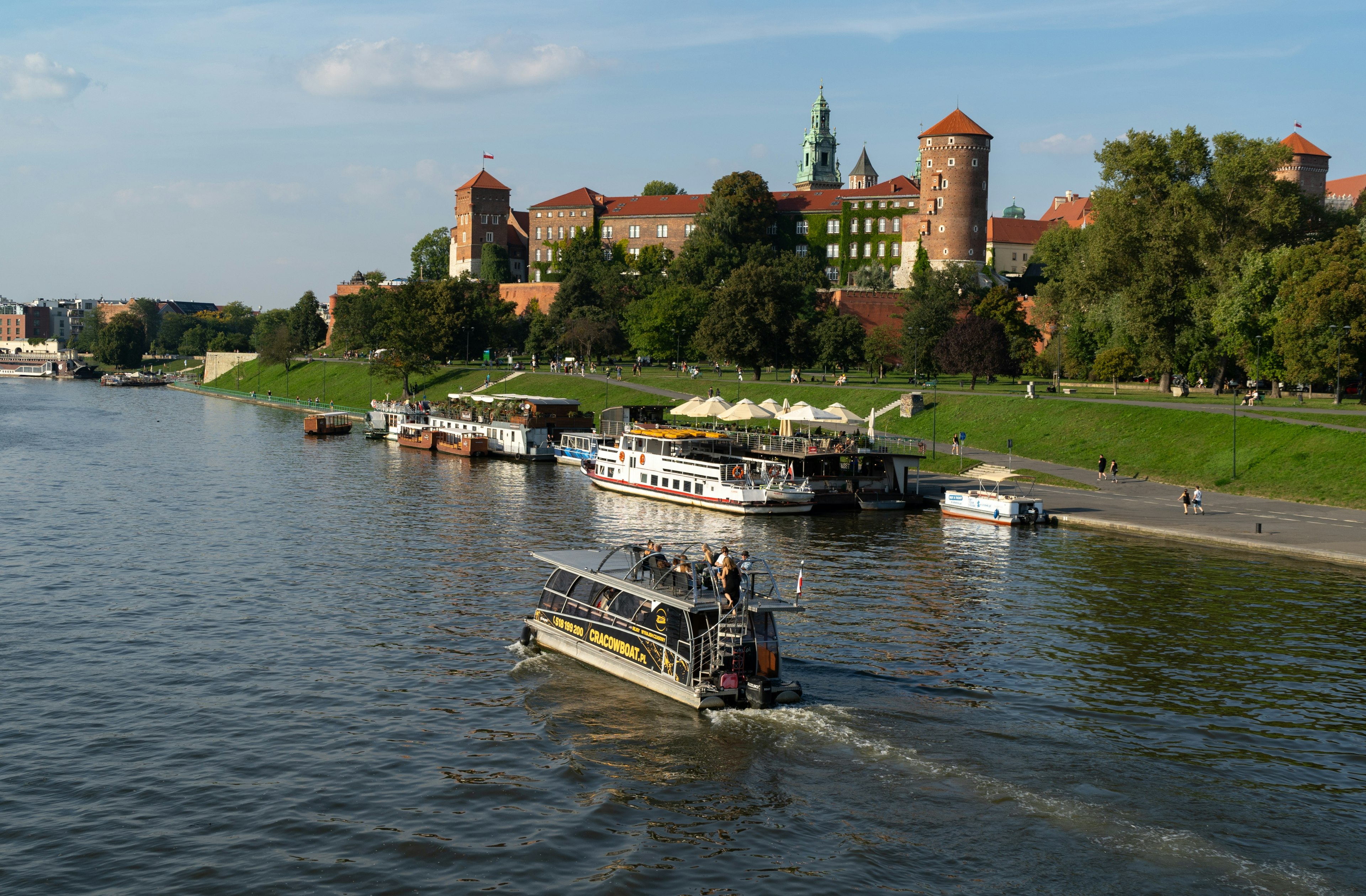 A boat cruises down a river, by other boats moored along the shore and a red-brick castle on a hill