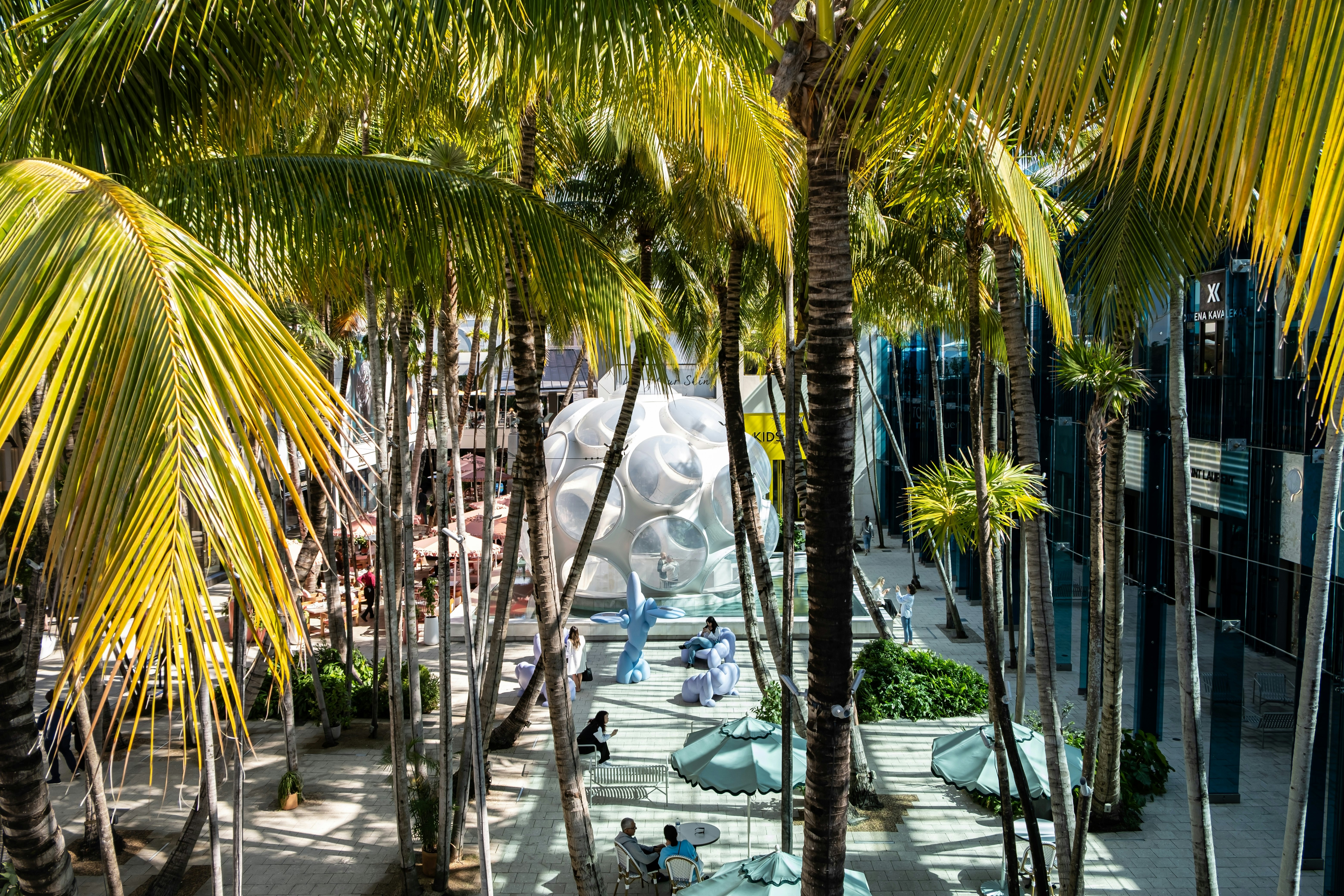 Palm trees loom over a courtyard with public art. People sit on benches and at tables relaxing.