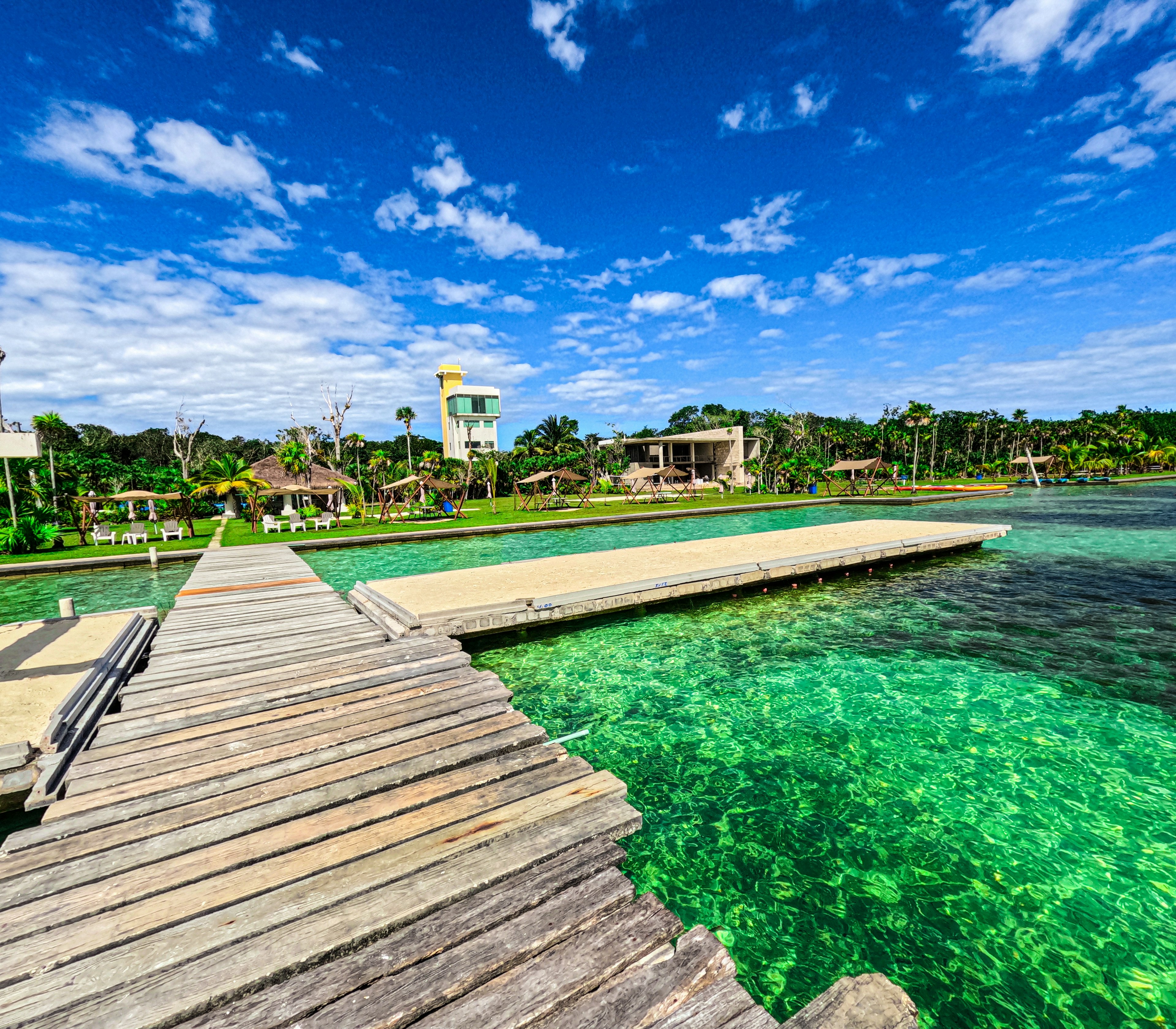 A boardwalk leads from a green and blue lake onto land where beach chairs and loungers sit in the shade