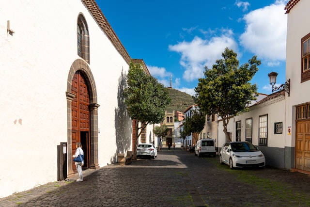 A woman stands in front of an elaborately carved door on a cobbled town streets lined with historic whitewashed buildings