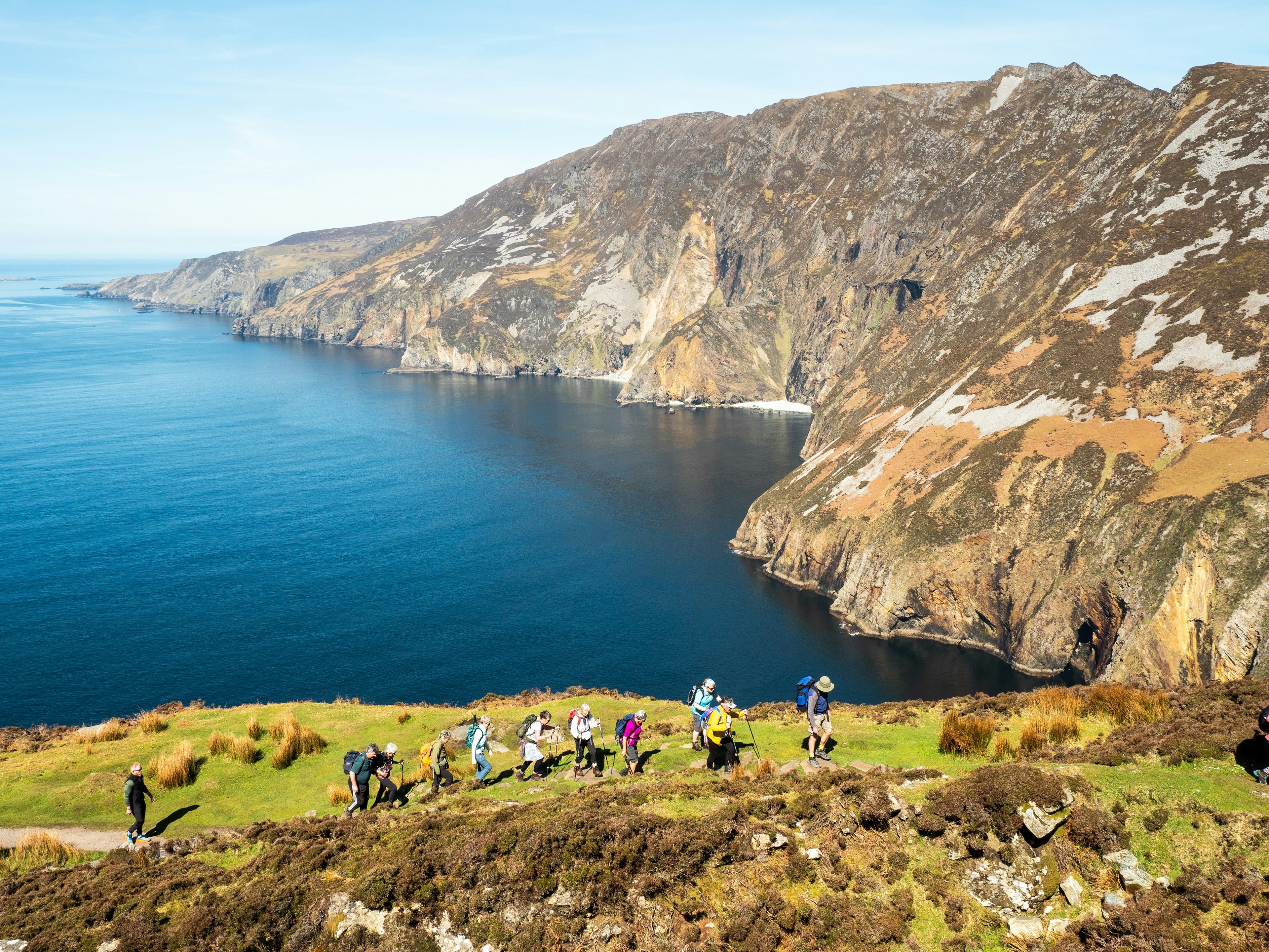 A group of hikers follow a narrow coastal path with sea cliffs rising nearby