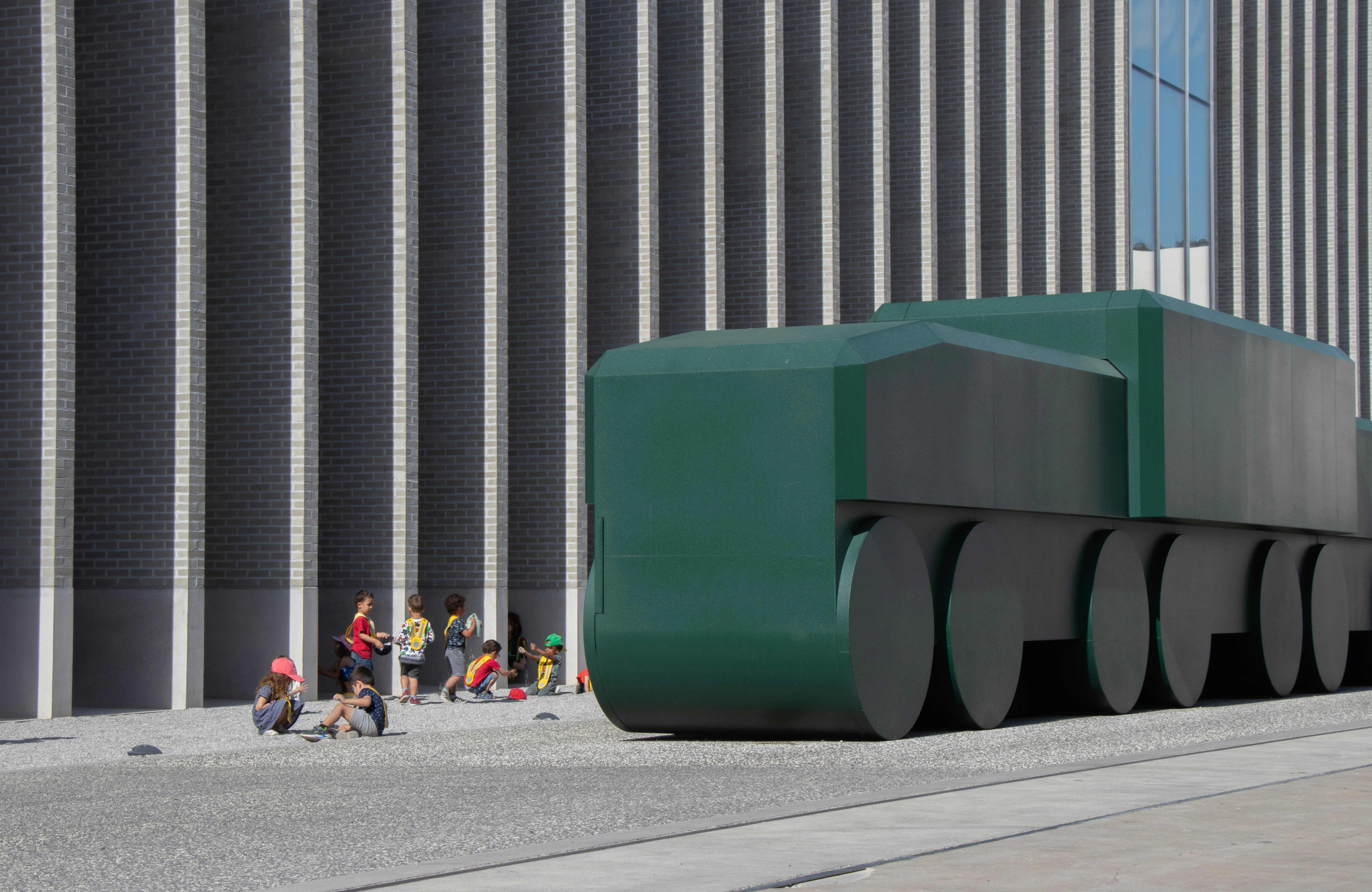 People gather in front of a large building with a large sculpture depicting a green, slightly abstracted green railcar