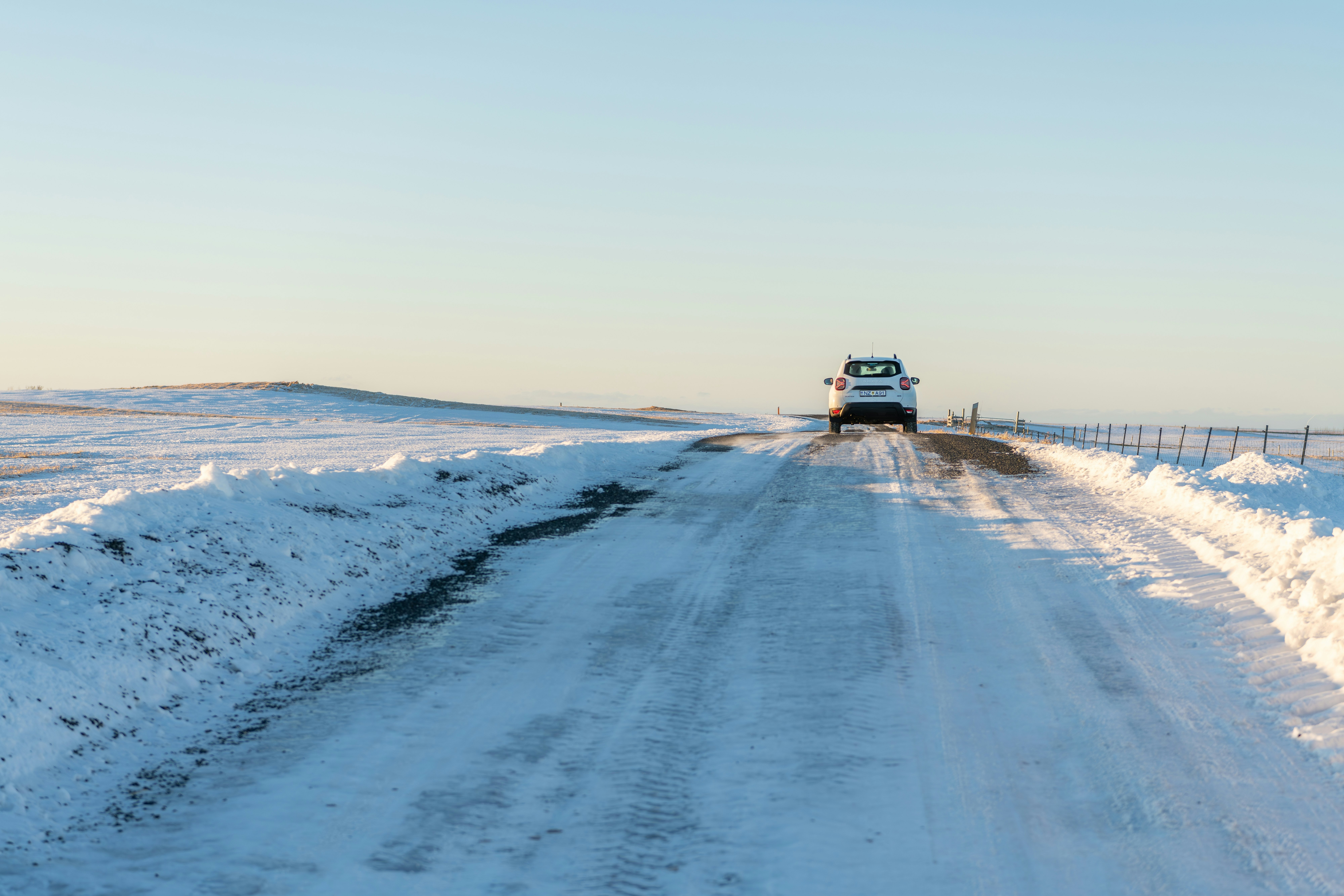 A rear view of a white Dacia Duster driving on a road covered with packed snow and ice through a snowy landscape