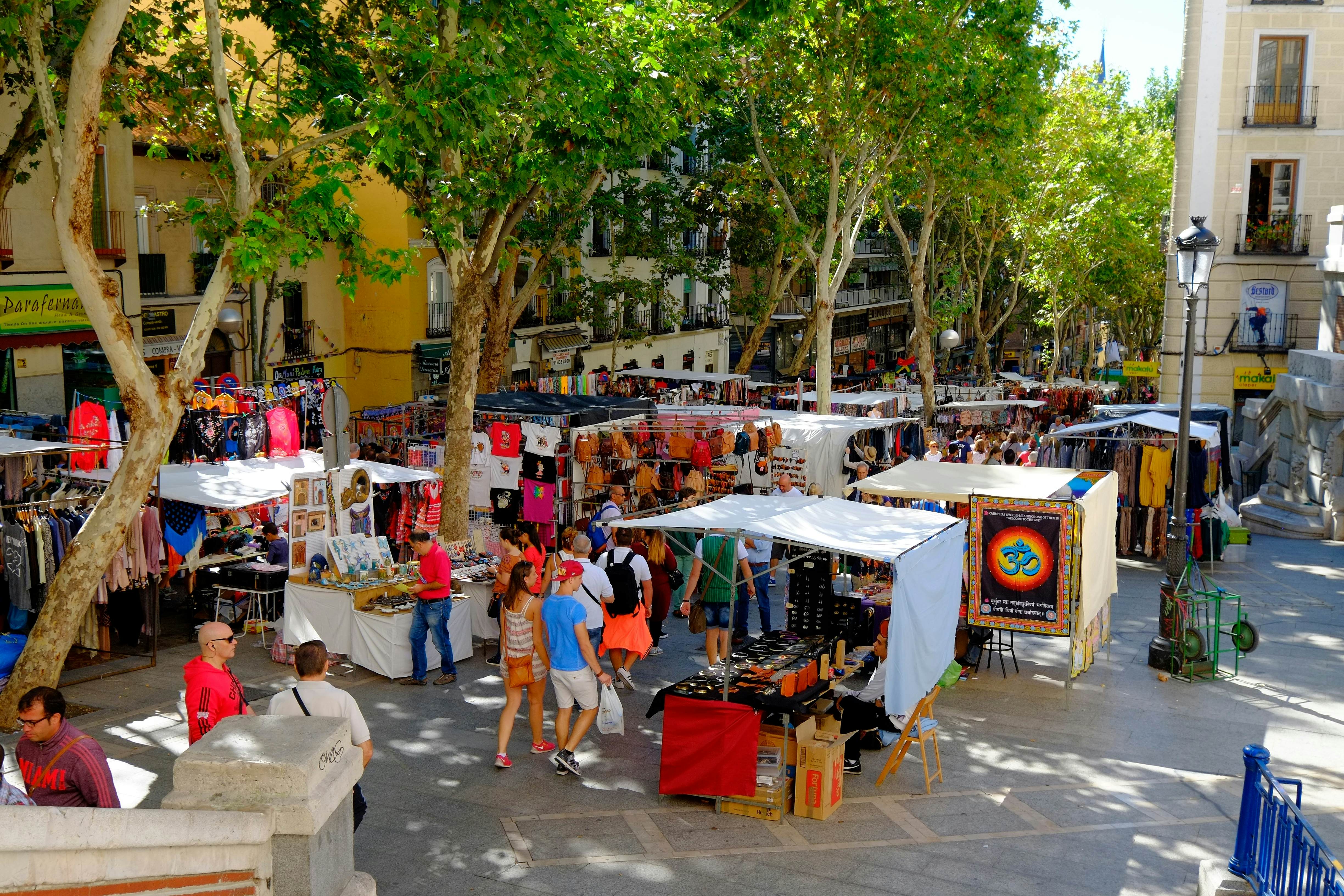 A tree-lined street hosts a vintage market on a sunny day, with lots of people buying and browsing