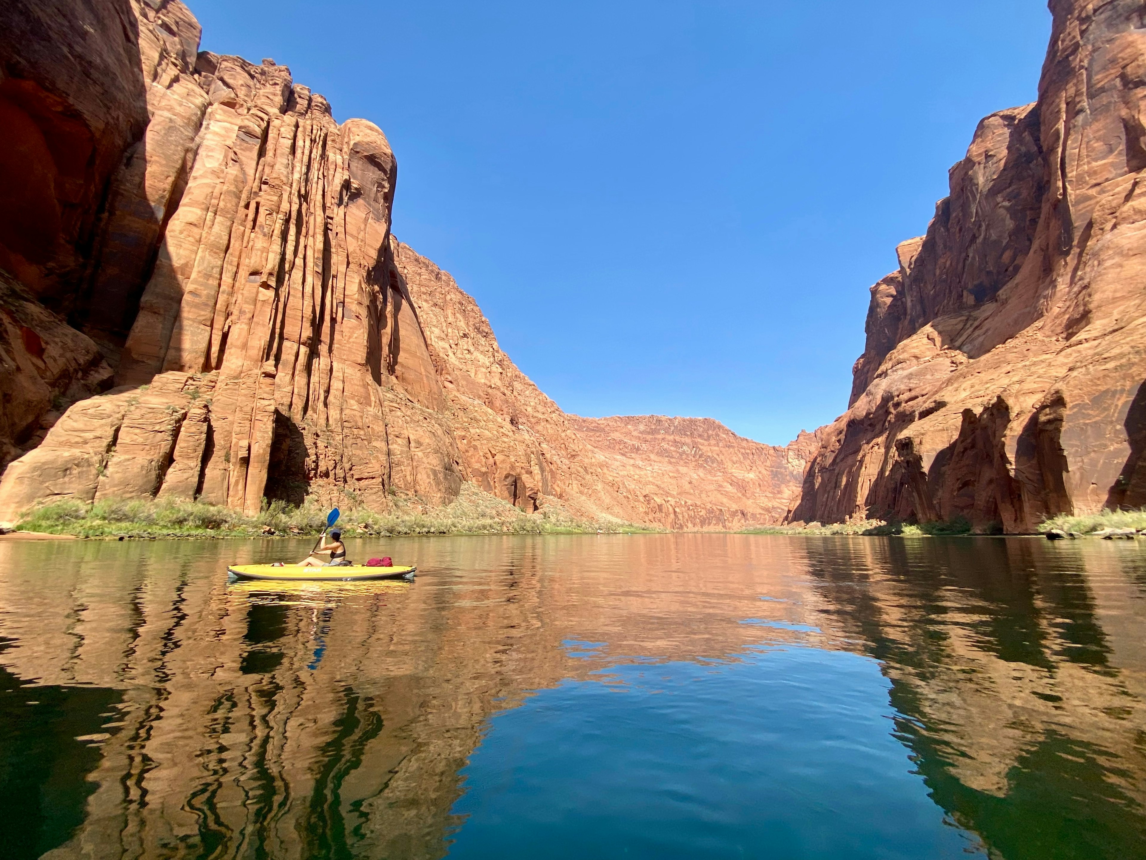 A woman paddles a yellow kayak on the Colorado River by the red-colored cliffs of Horseshoe Bend, Arizona