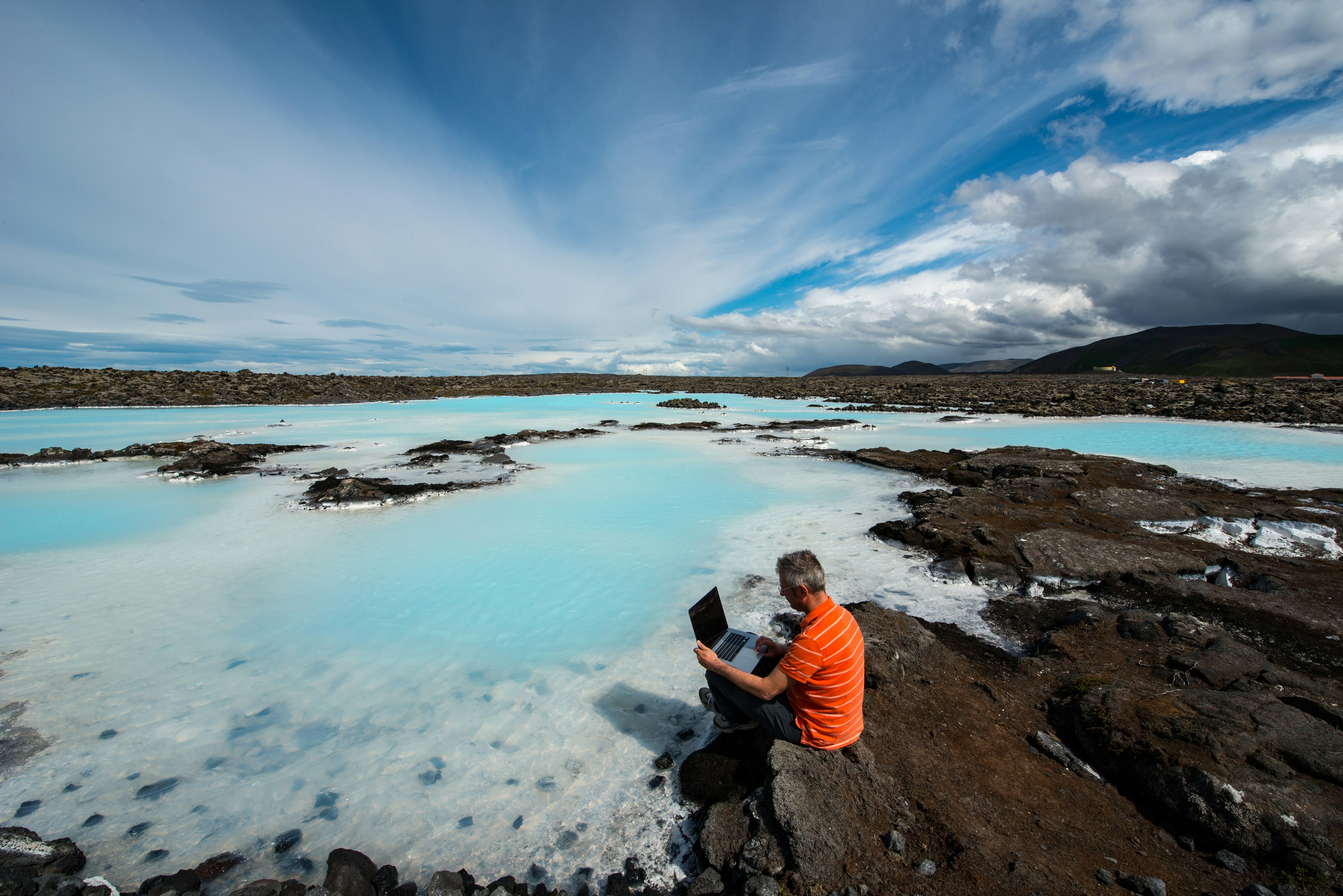 A man in an orange shirt works on a laptop while sitting on a rock at an inlet in Iceland