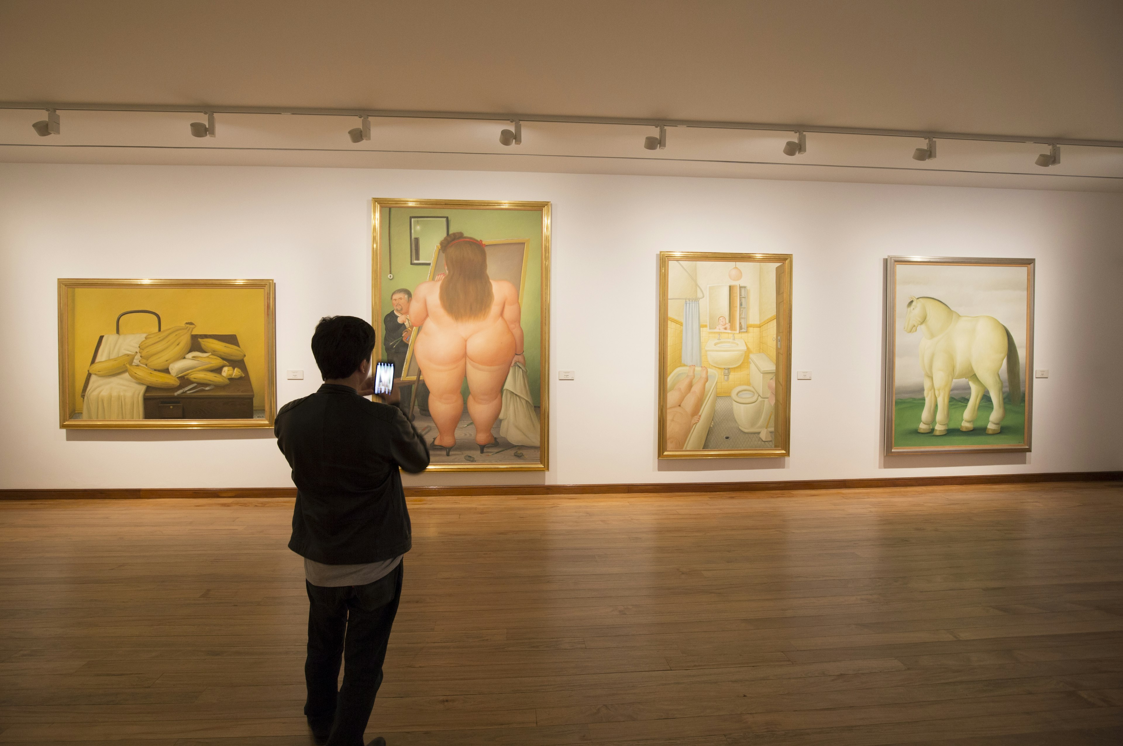 A person stands art gallery taking a picture of paintings of models and still-life subjects