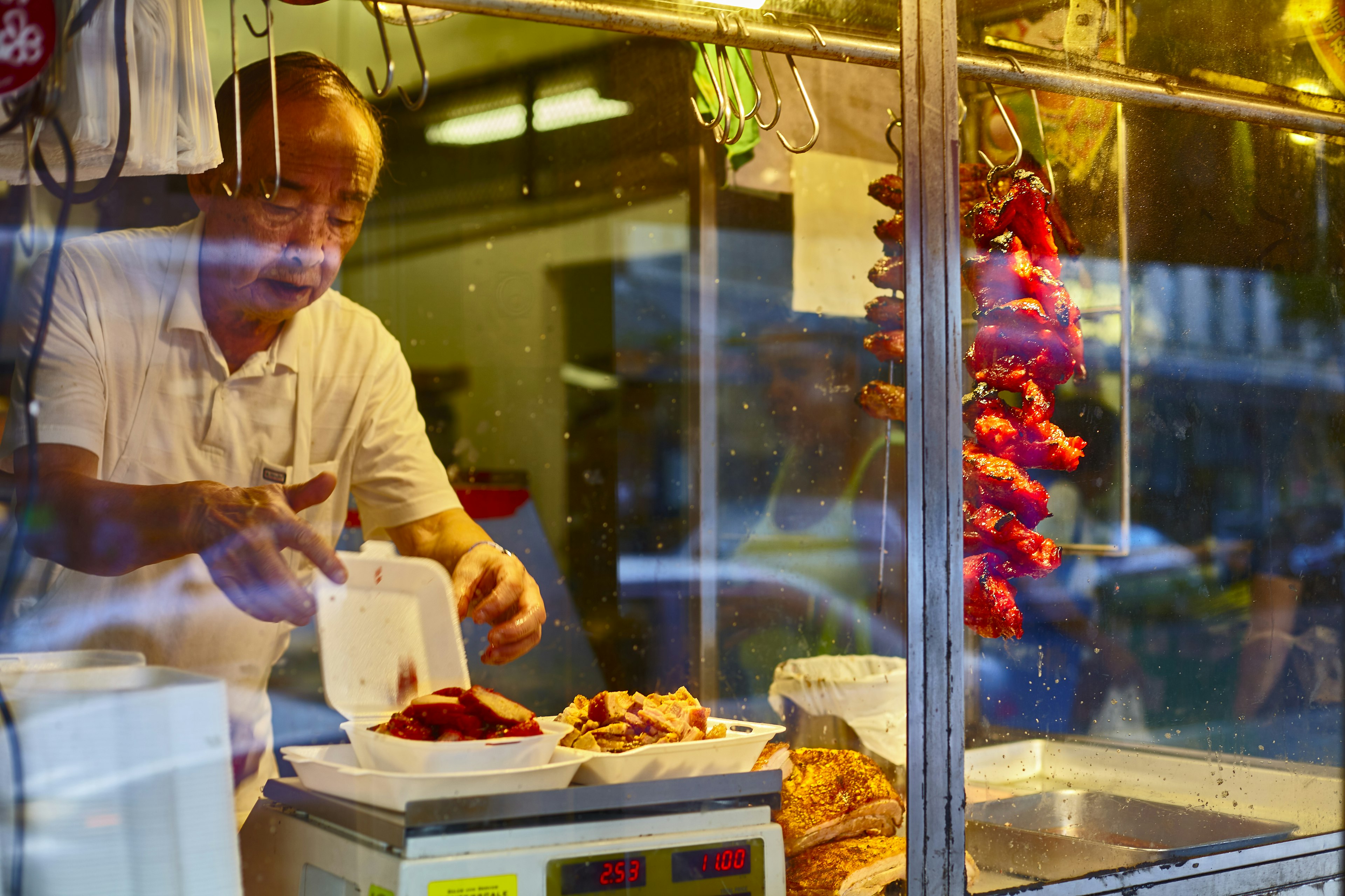 A man prepares roast pork for a customer behind a glass wall at night in the Chinatown Historic District, Honolulu, Hawaii, USA