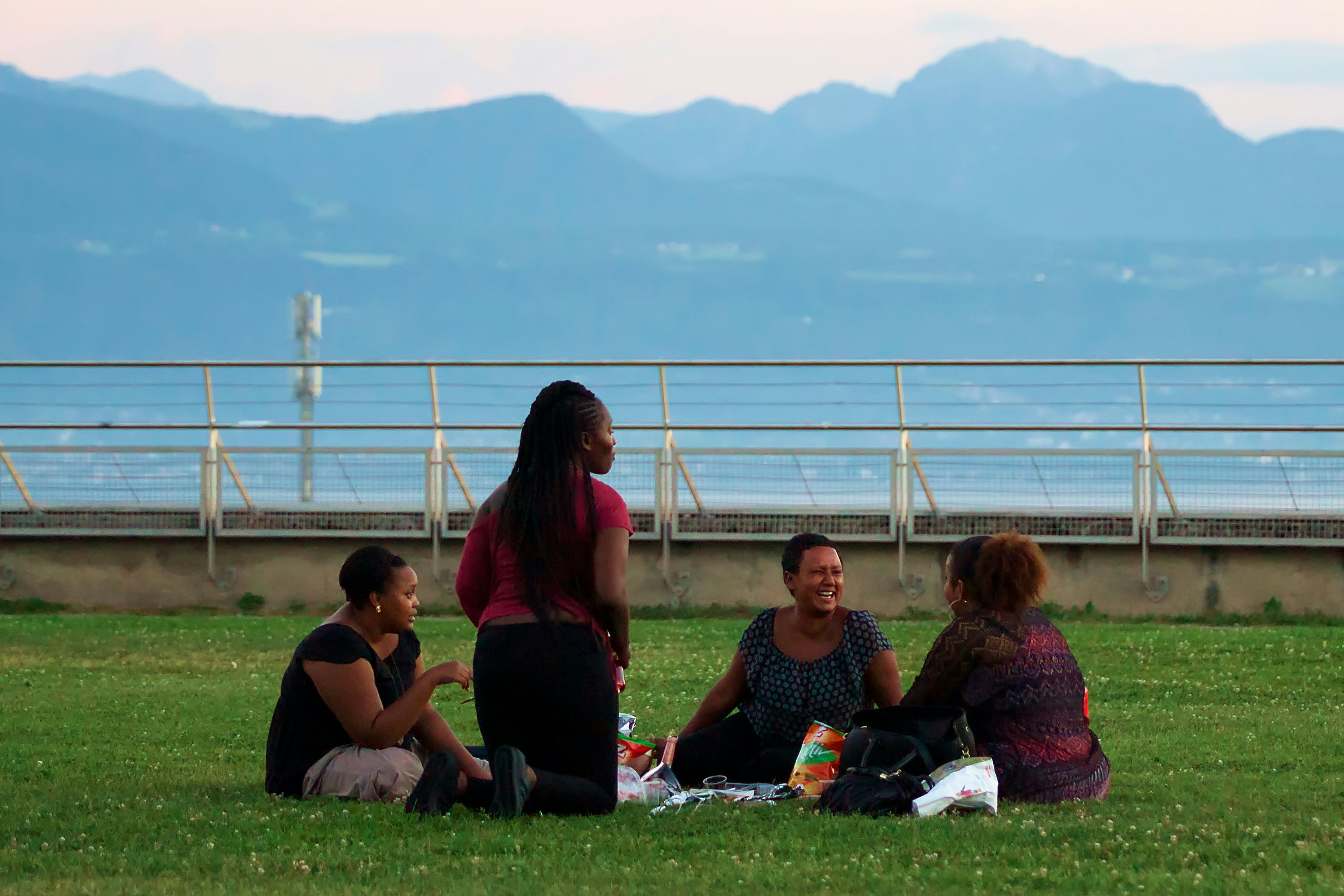 A group of four women enjoys a picnic on a green lawn in a park overlooking mountains