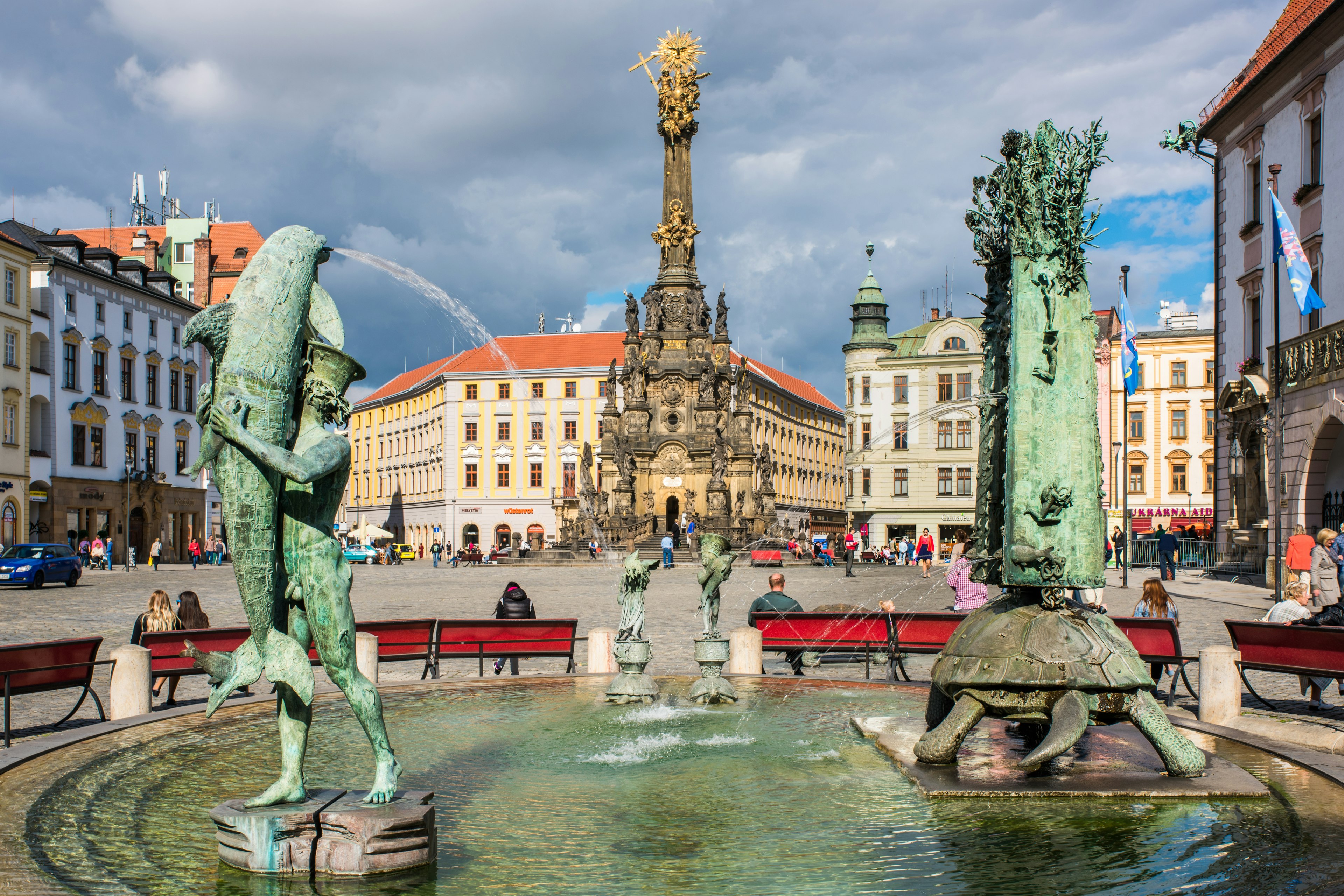 An elaborately carved column is seen beyond two sculptural fountains in a city square