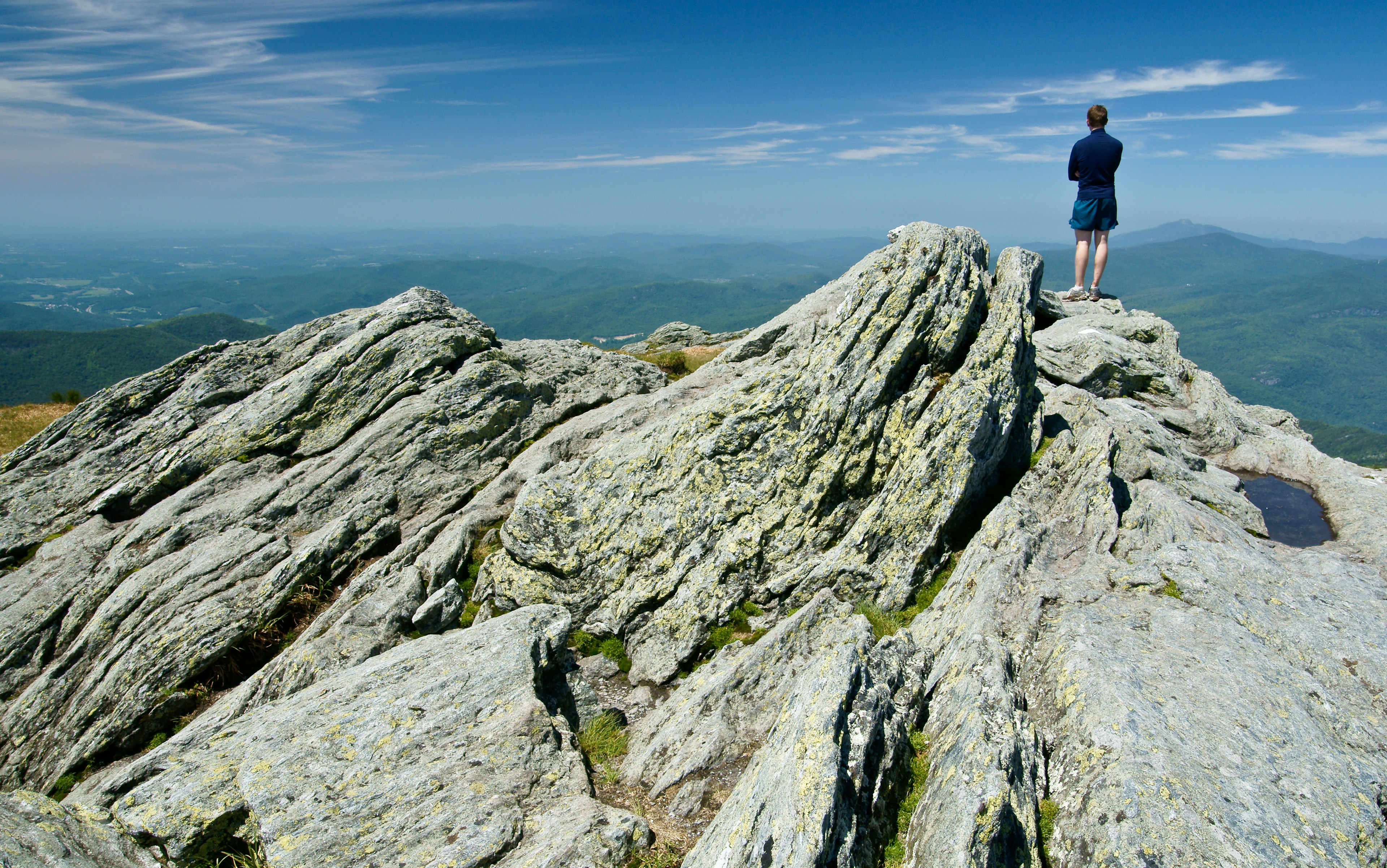 A solo hiker stands on the peak of some rocks looking down at the view below