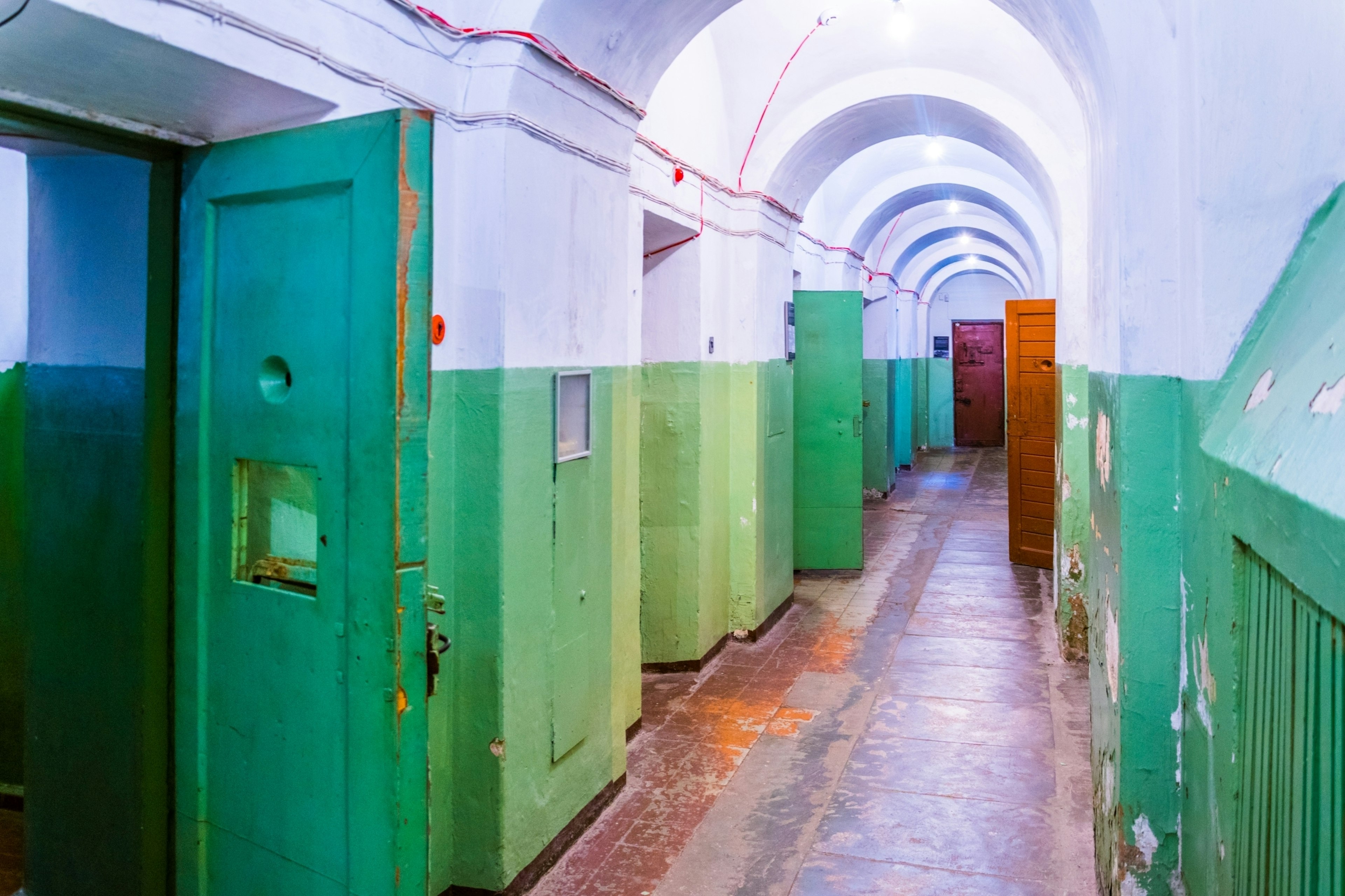 A basement corridor painted green and white with doors open to individual prison cells