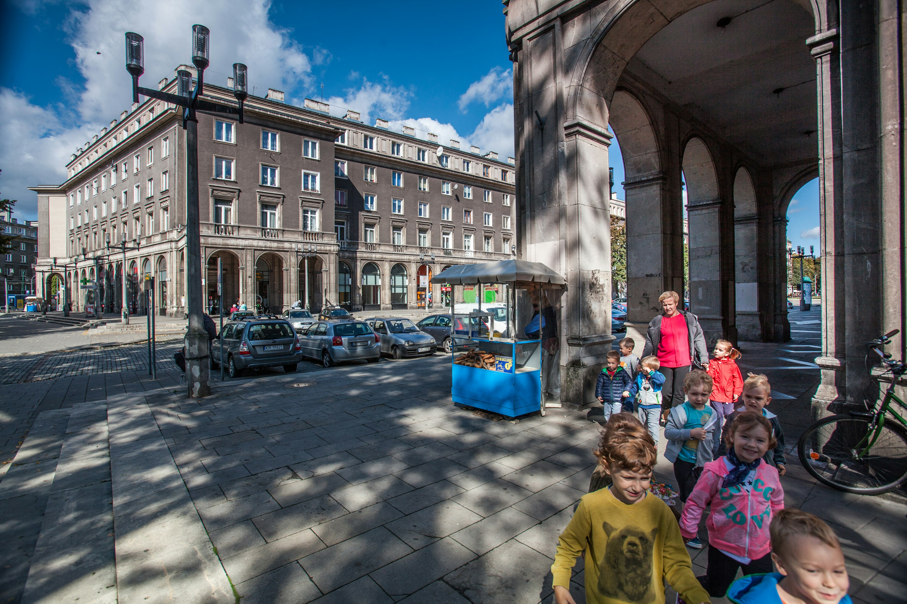 Children walk through a colonnade in the shadows from a tree against a streetscape of gray apartment builidings