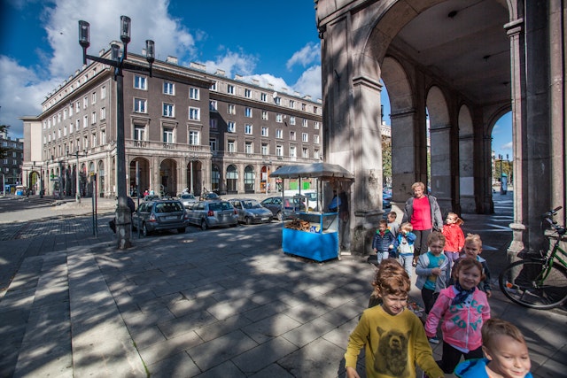 Children walk through a colonnade in the shadows from a tree against a streetscape of gray apartment builidings