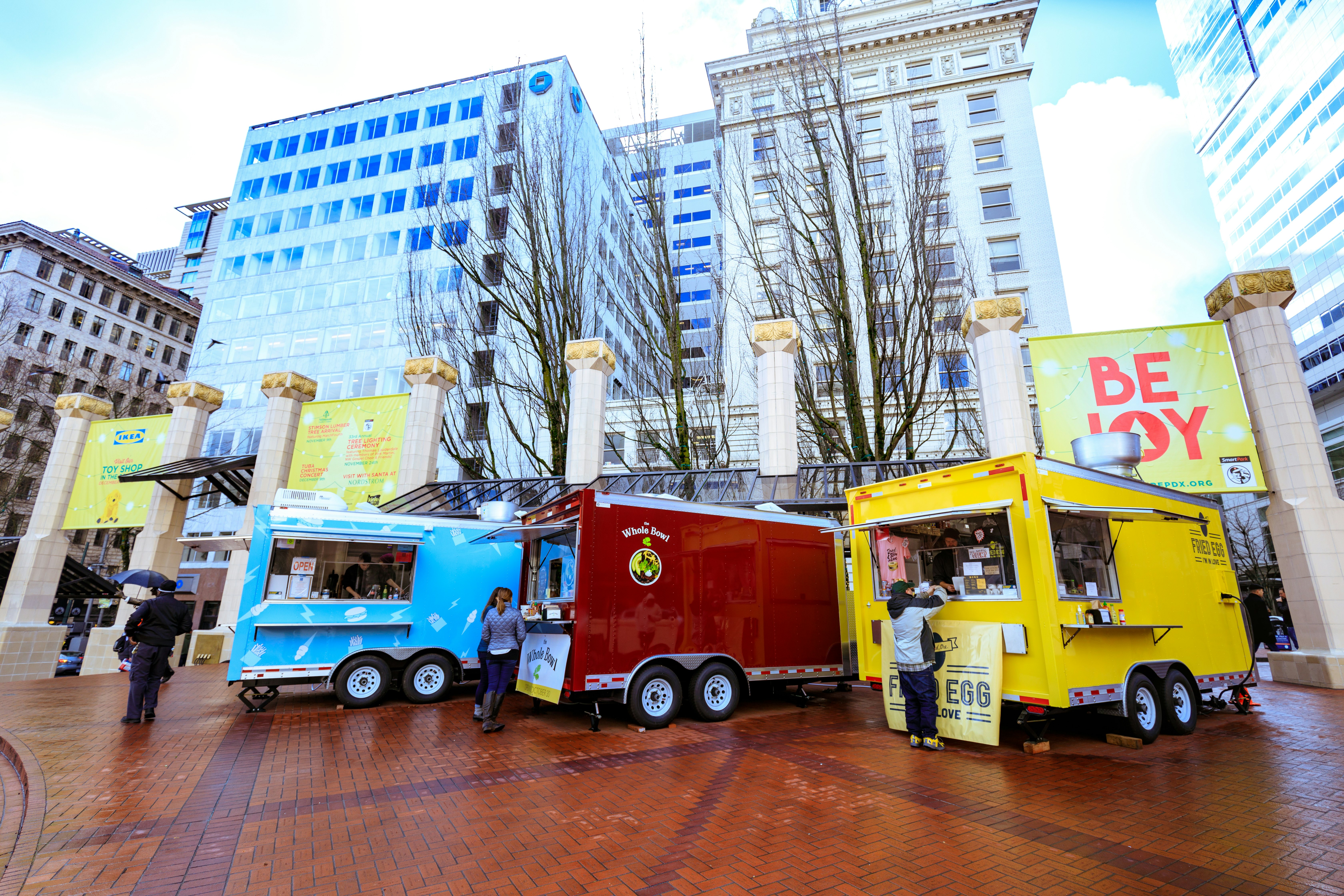 Three brightly colored food trucks parked in Pioneer Courthouse Square on a rainy day, surrounded by office buildings, Portland, Oregon, USA