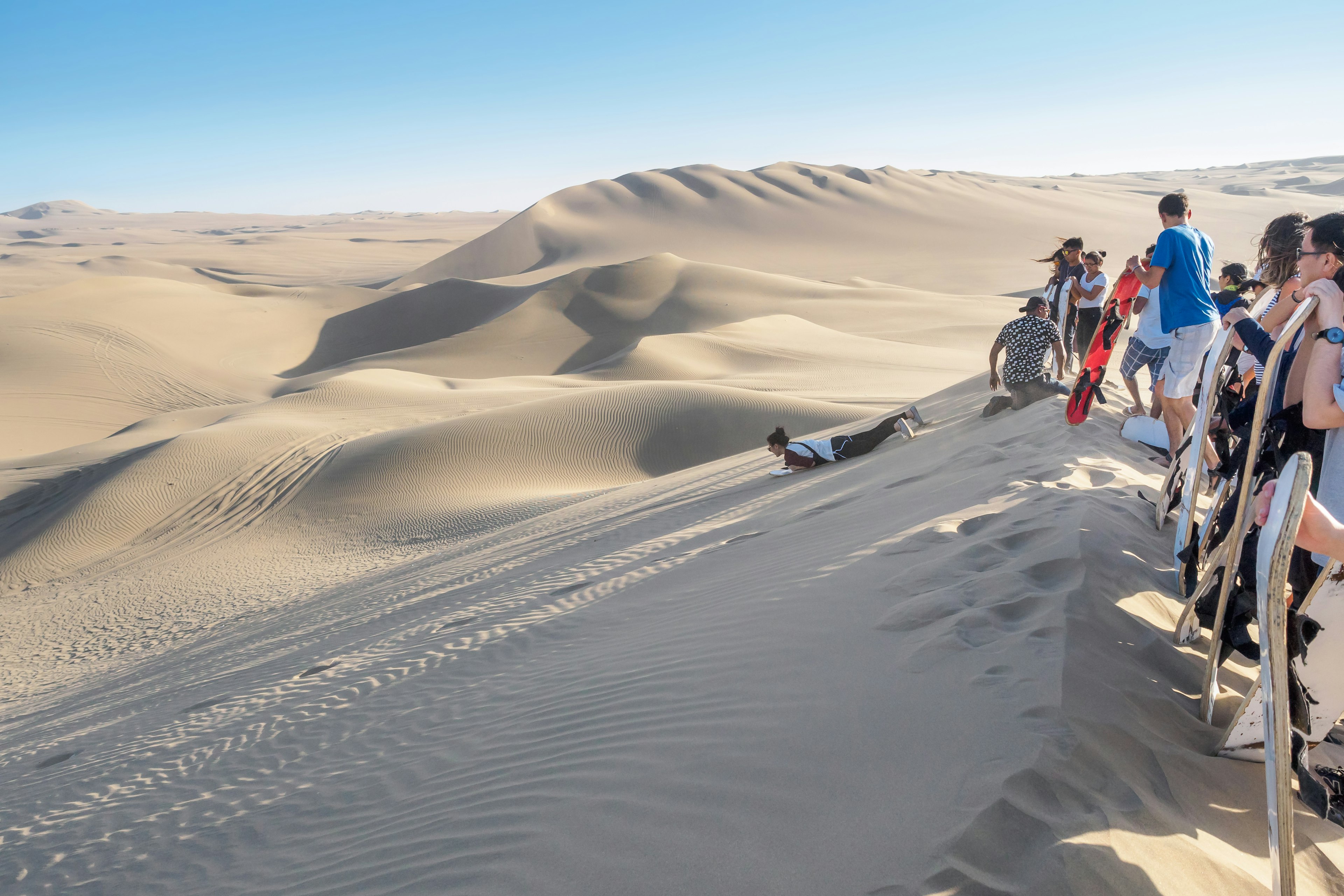 Tourists stand at the ridge of a massive sand dune as they wait to go sandboarding in the desert