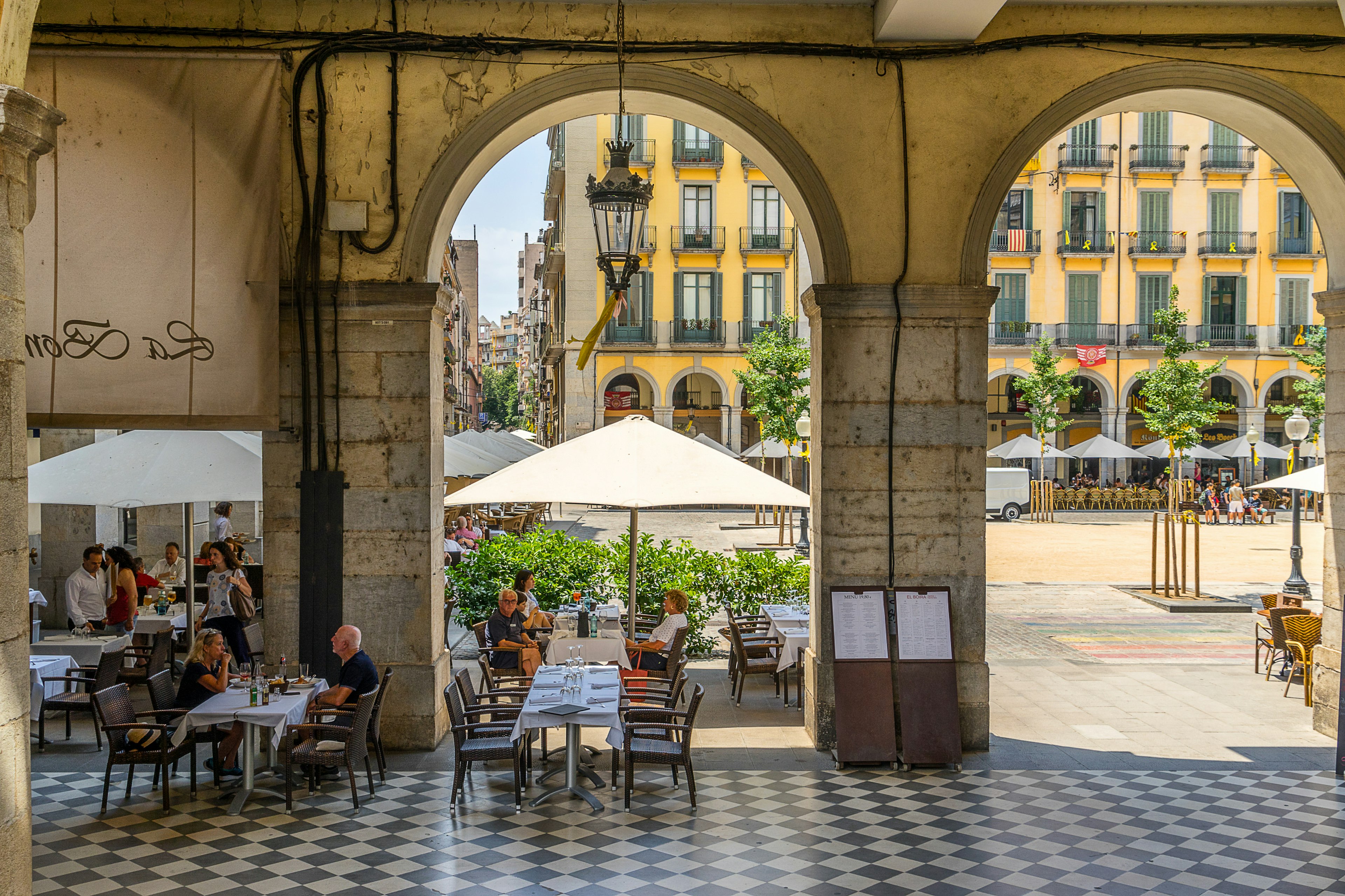 People seated at a restaurant in the shade framed by some archways on the edge of a city square